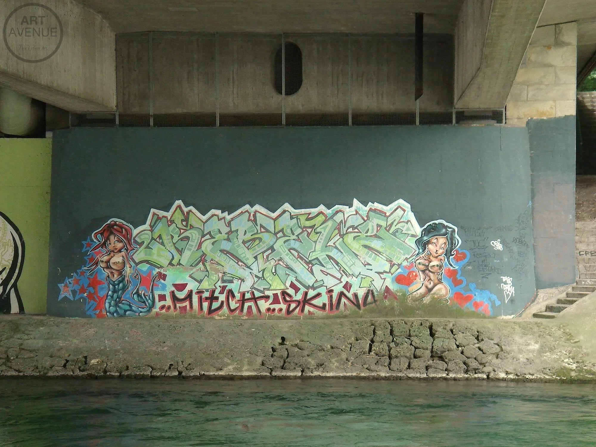 Graffiti Kunst an einer Wand, mit zwei cartoonartigen Meerjungfrauen auf beiden Seiten, einer mit roten Haaren und einer mit schwarzen Haaren, sowie dem Namen "MECHSKINA" in der Mitte langsam.