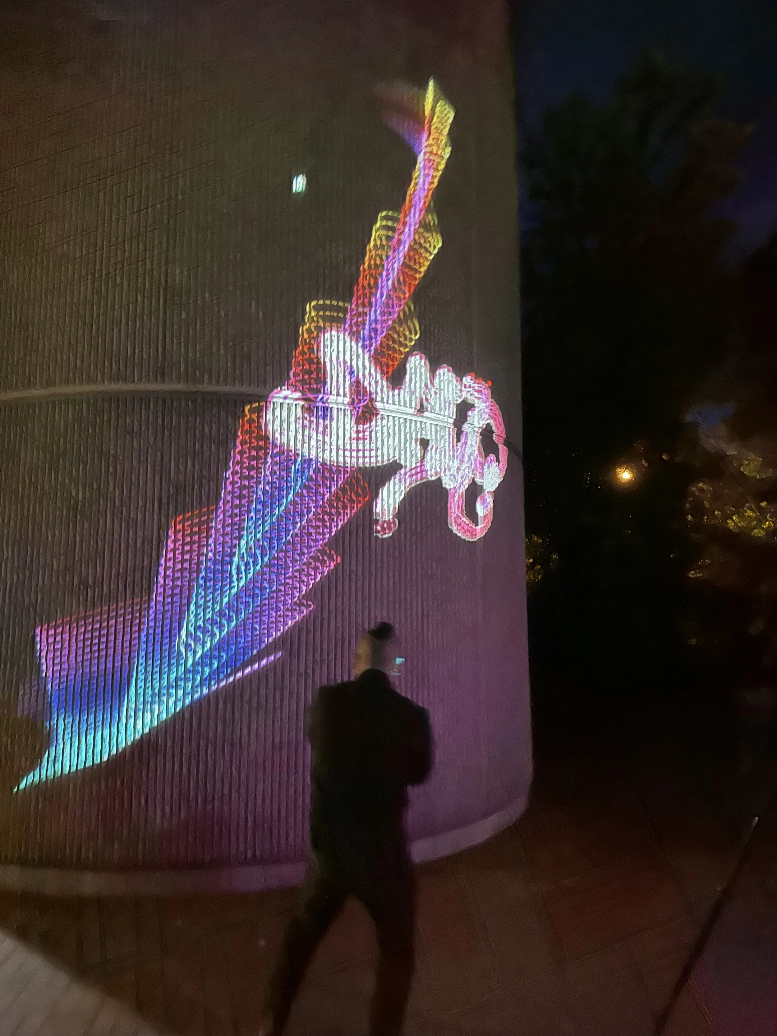 Eine nächtliche Aufnahme eines Gebäudes mit Lichtprojektion. Auf der Wand wird eine bunte, digitale Kunstprojektion eines Motorrads und eines Mopeds angezeigt.