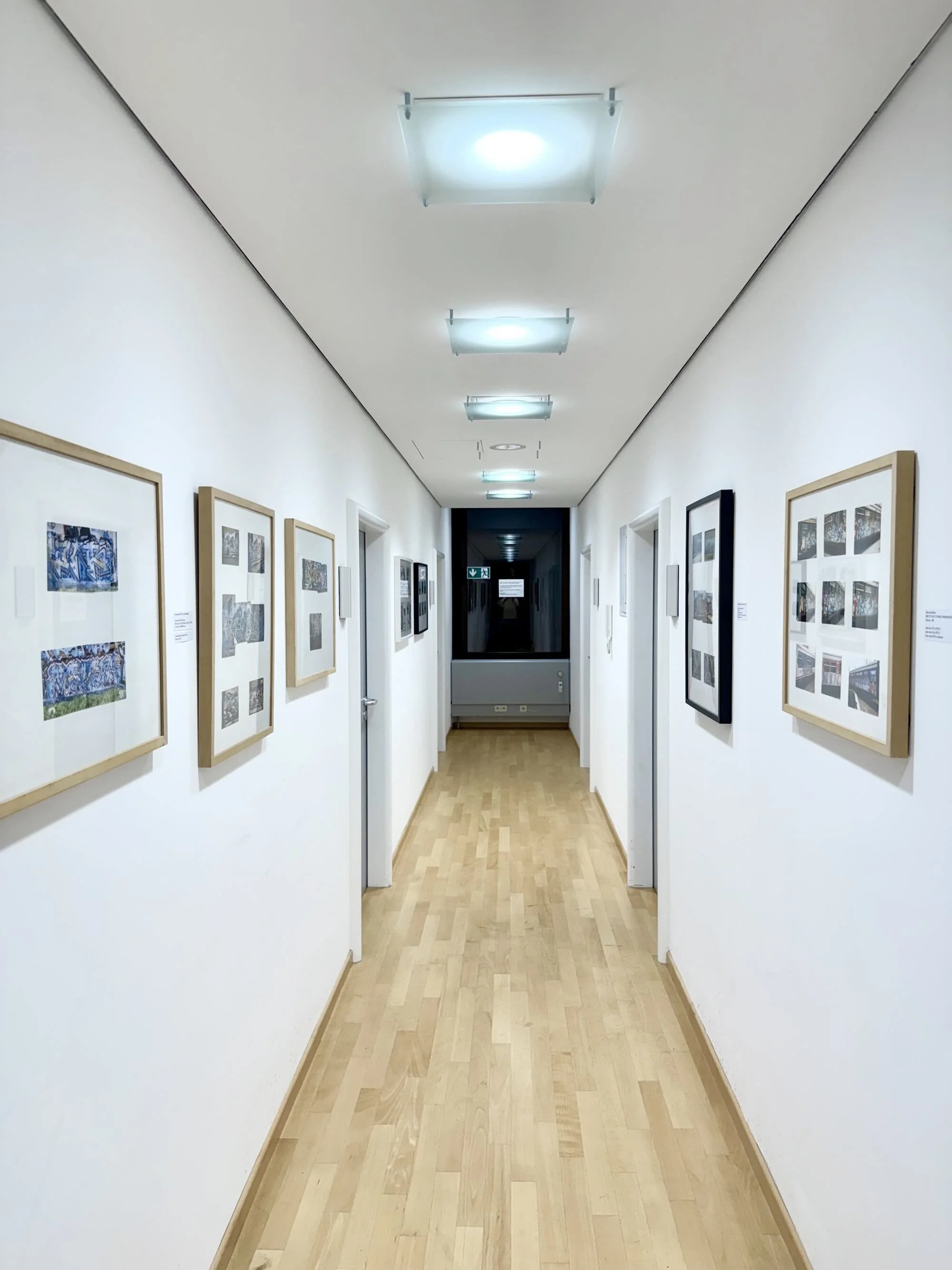 Ein schmaler Flur in einer Kunstgalerie mit weißen Wänden, an denen gerahmte Kunstwerke hängen, und einem Holzboden. Deckenleuchten sind gleichmäßig verteilt.