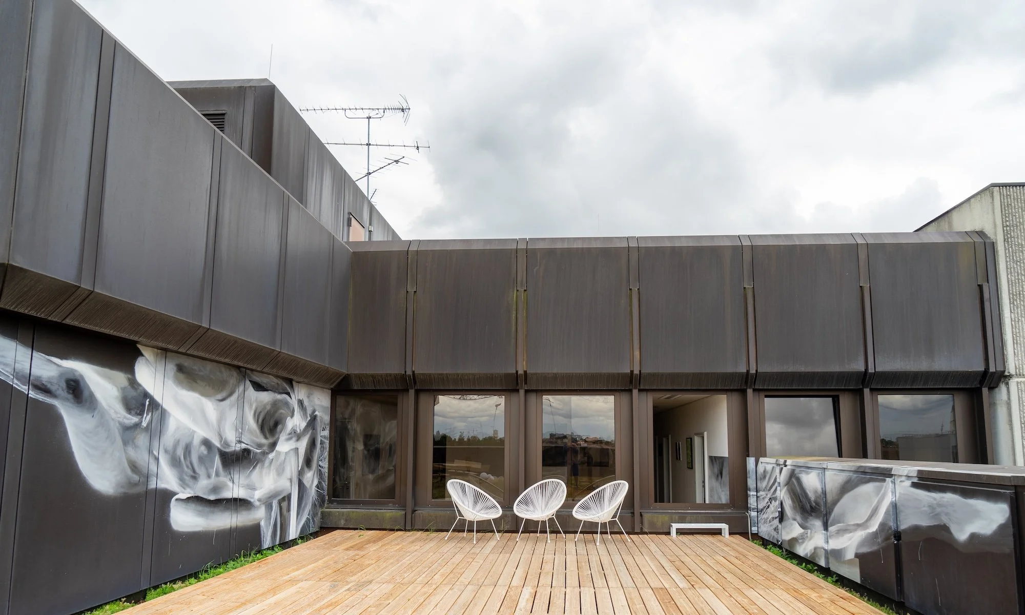Modernes Gebäude mit grauer Metallfassade, Balkone und einer Terrasse mit drei weißen Stühlen im Freien, bei bewölktem Himmel.