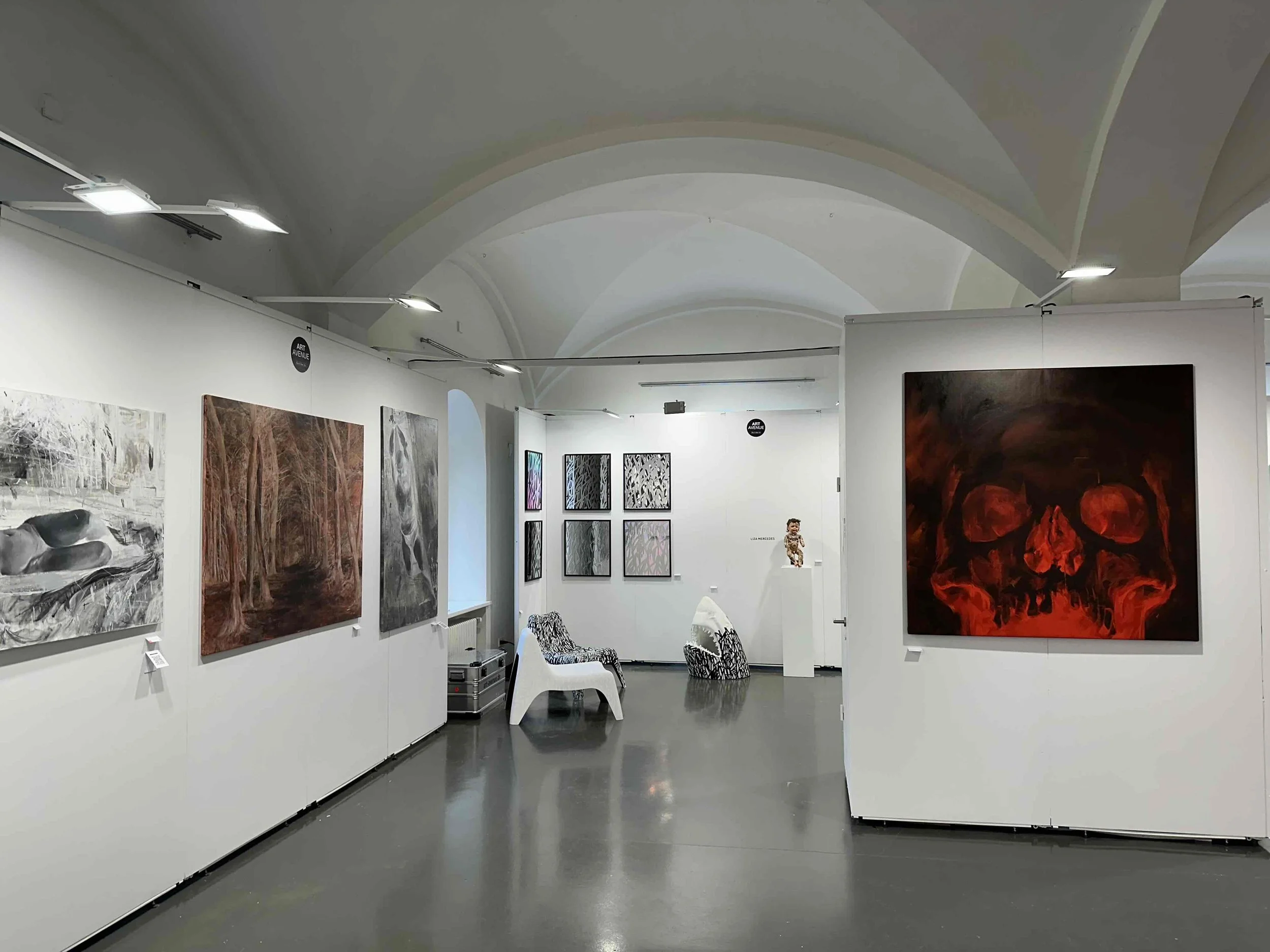 Innenraum einer Galerie mit weißen Wänden, auf denen mehrere Kunstwerke hängen, darunter Gemälde mit Natur- und Tiermotiven. Es sind weiße Sitzgelegenheiten und eine Skulptur im Hintergrund zu sehen.