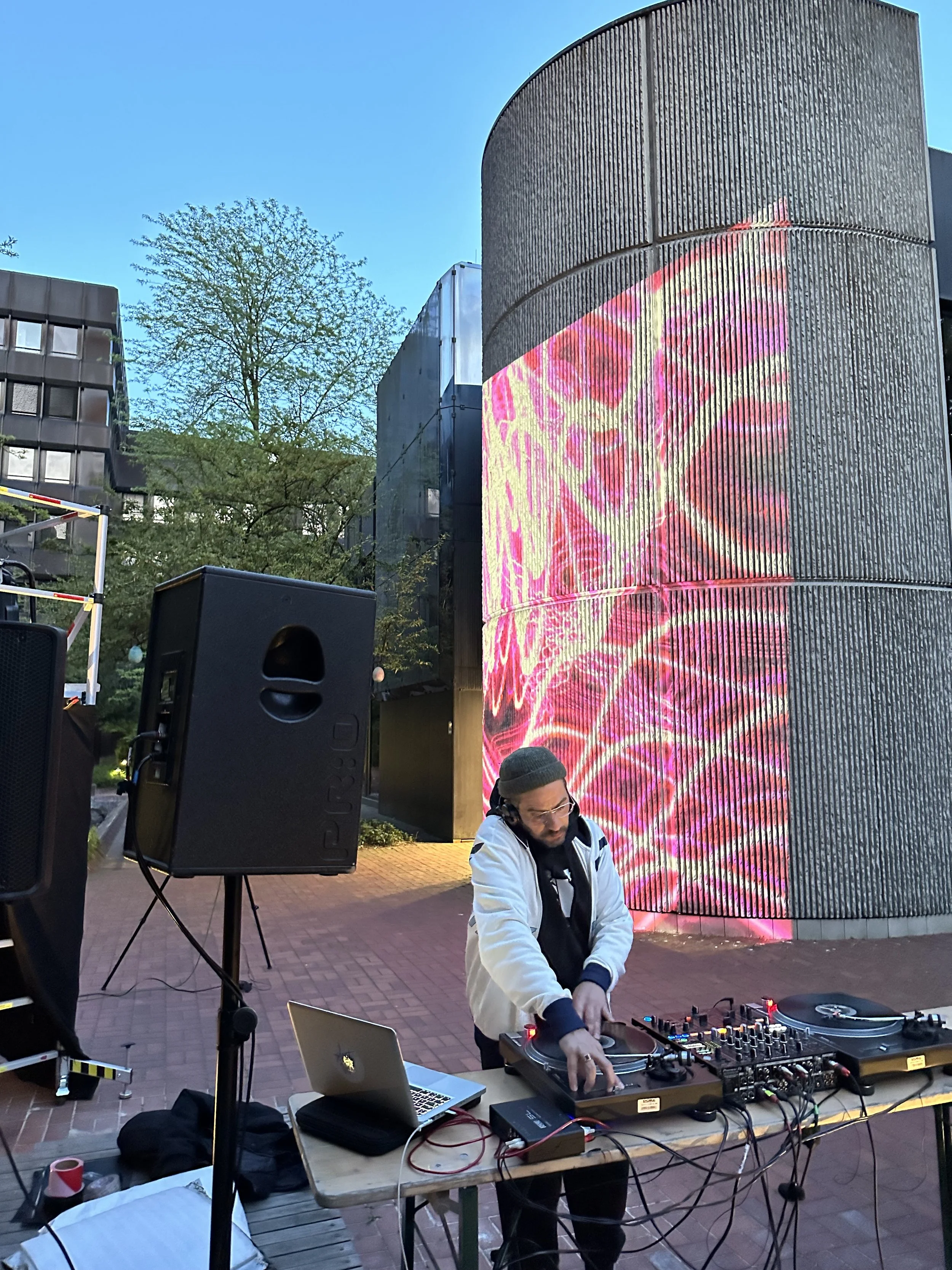 DJ spielt an einem offenen Plattenteller vor einem hell erleuchteten, runden Gebäude mit digitaler Projektion auf der Fassade.