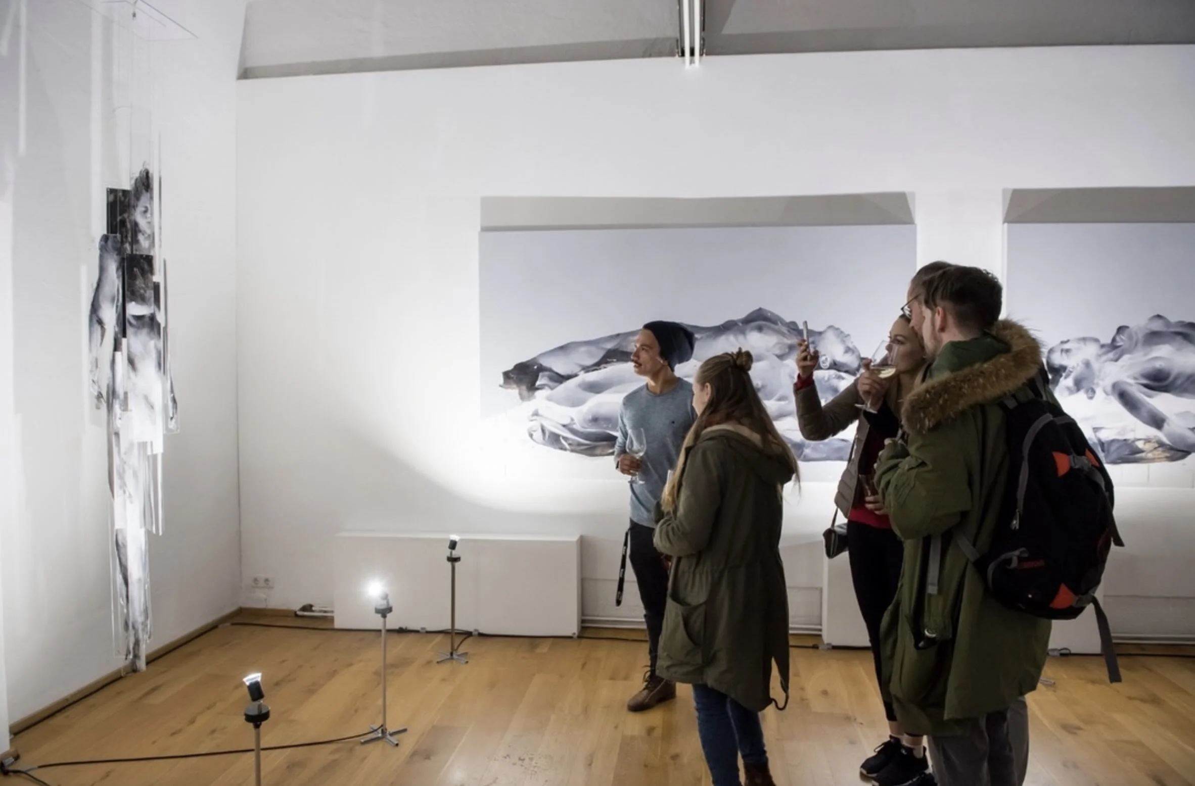 Vier Menschen in einer Kunstgalerie betrachten und fotografieren Kunstwerke an weißen Wänden, die Minikameras auf Ständern beleuchten und eine große Wandmalerei zeigen.