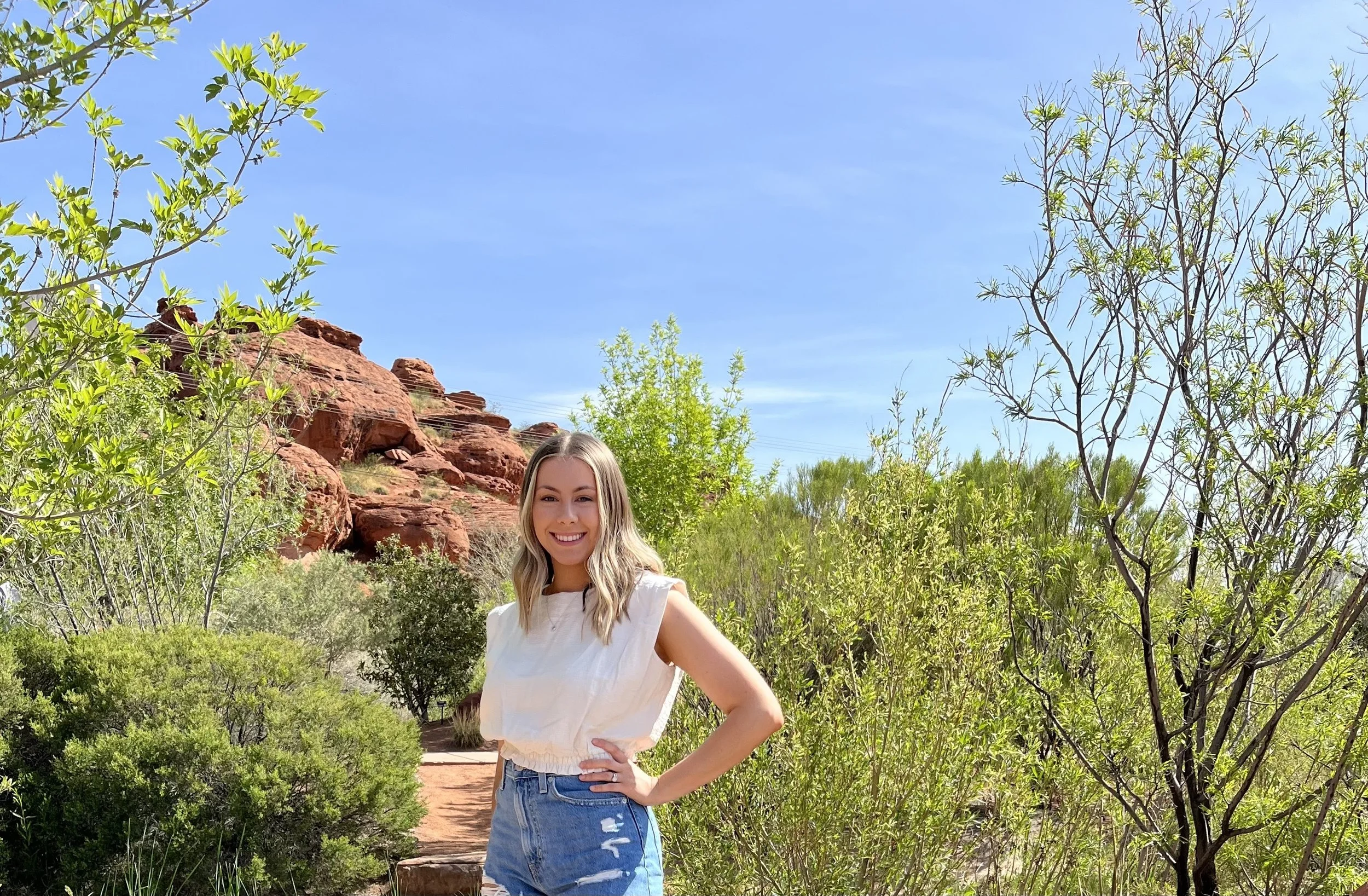 A smiling woman with blonde hair wearing a sleeveless white top and distressed denim shorts stands in a desert landscape with red rocks, green shrubs, and trees under a clear blue sky.