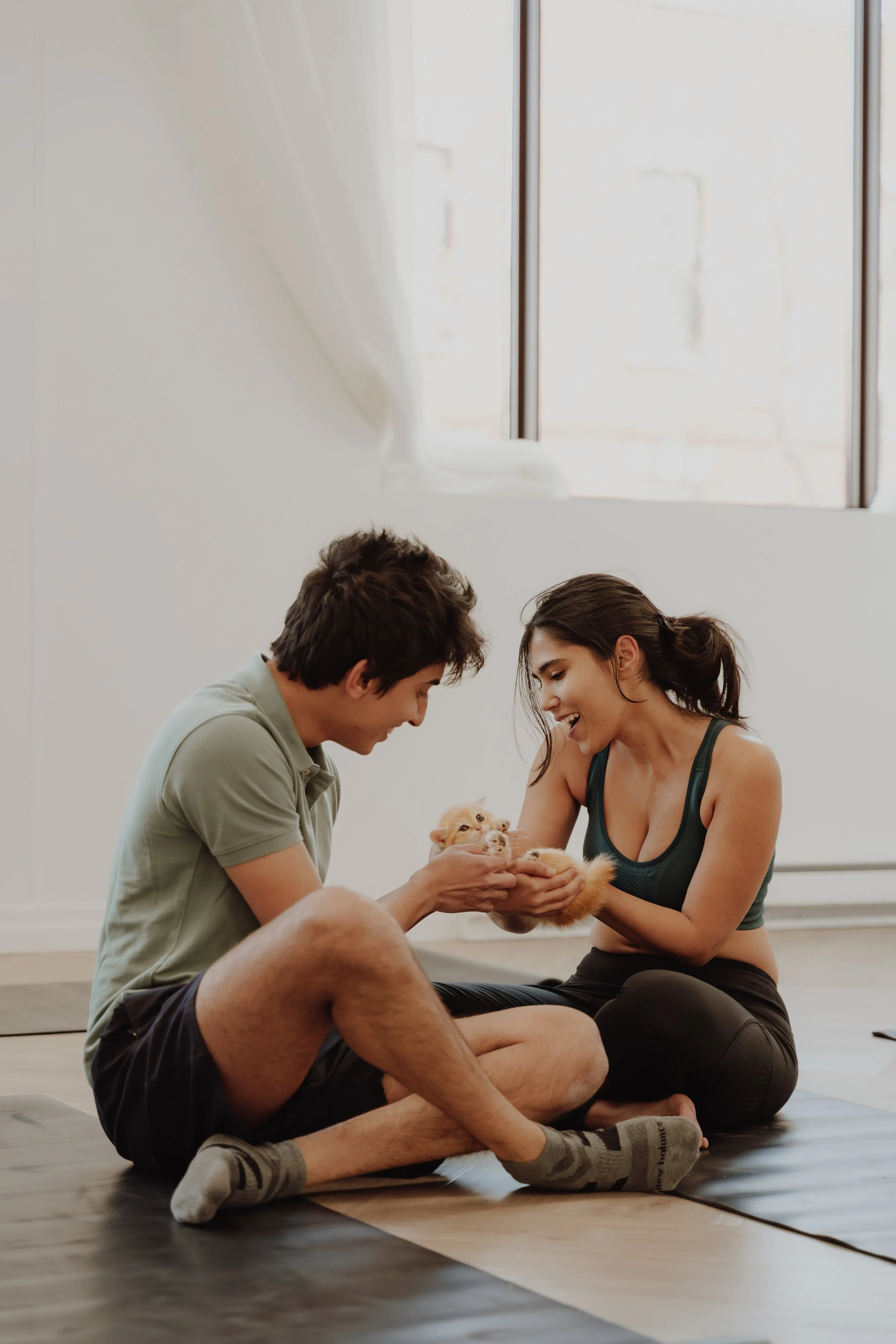 A young man and woman are sitting on yoga mats on the floor, playing with a small puppy inside a bright room with large windows.