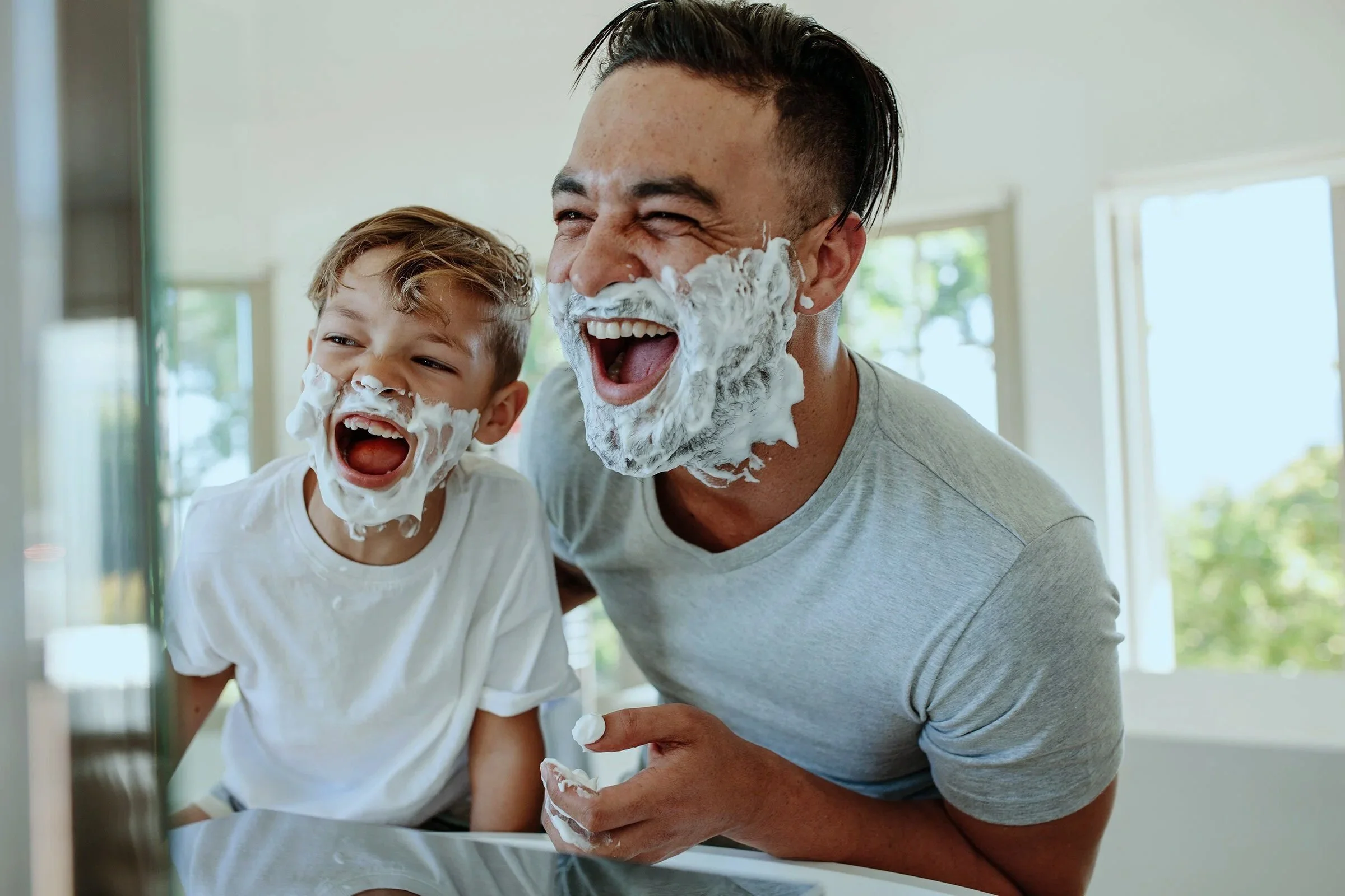 A man and a young boy with shaving cream on their faces, laughing and having fun together in a bright, sunny room.