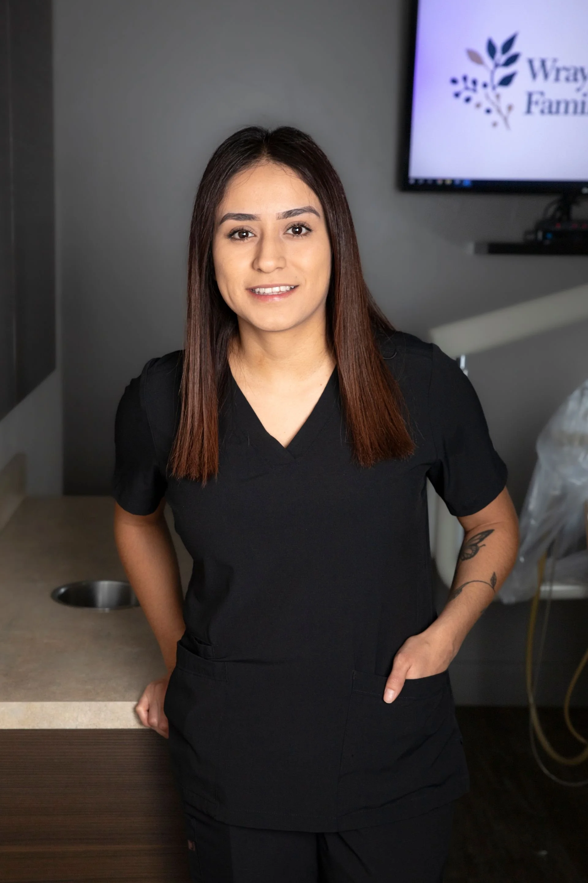 A woman in black medical scrubs standing in an indoor setting with a desk and a monitor in the background.