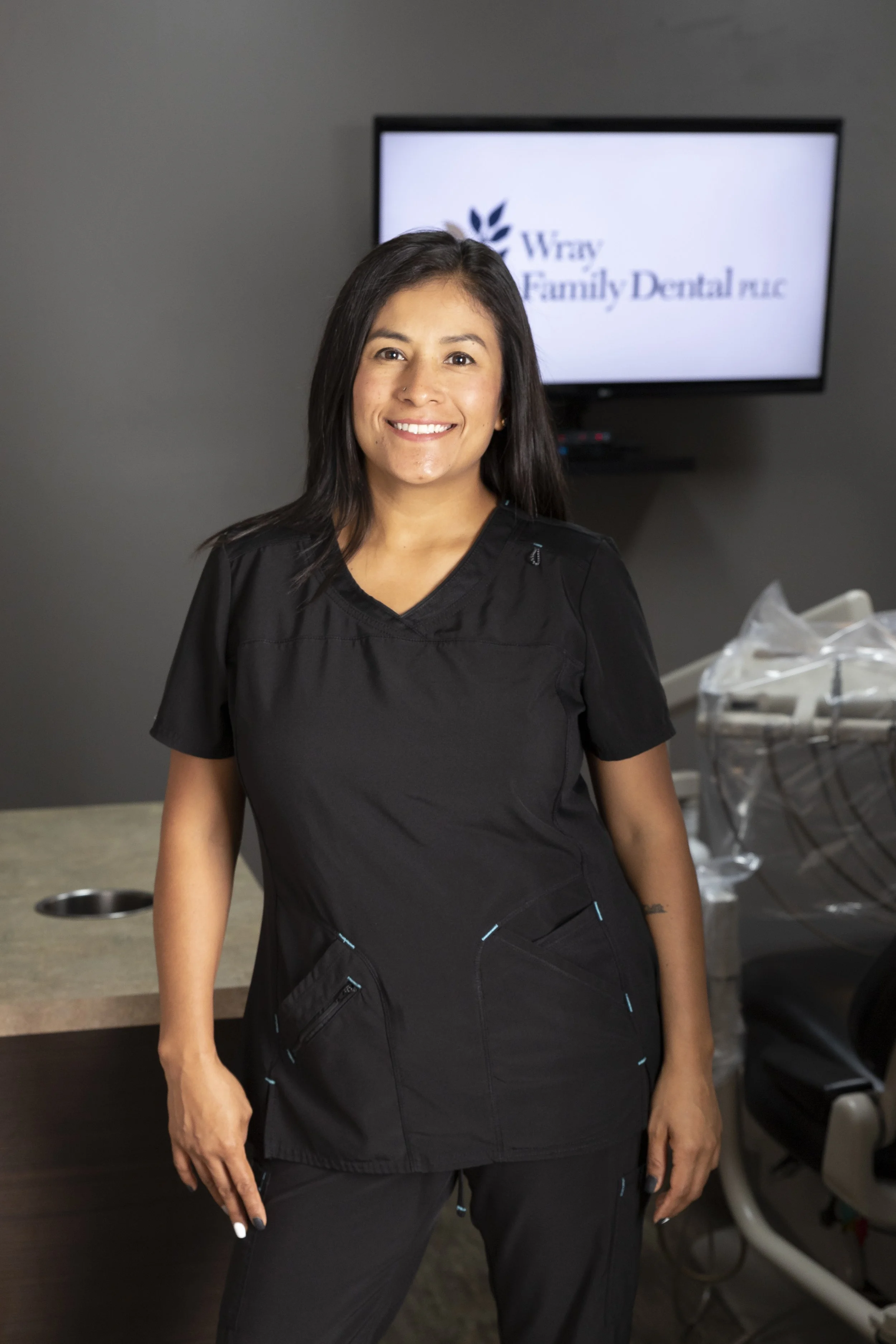 A smiling woman in black scrubs standing in a dental office