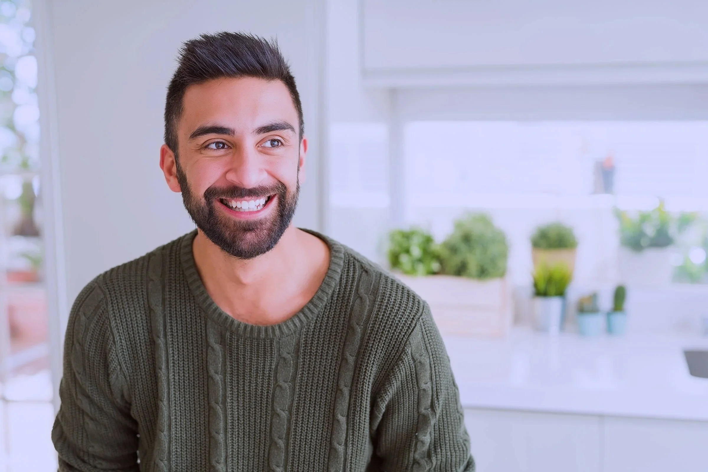 A smiling man with dark hair and a beard, wearing a green cable-knit sweater, sitting indoors with a bright window and green potted plants in the background.