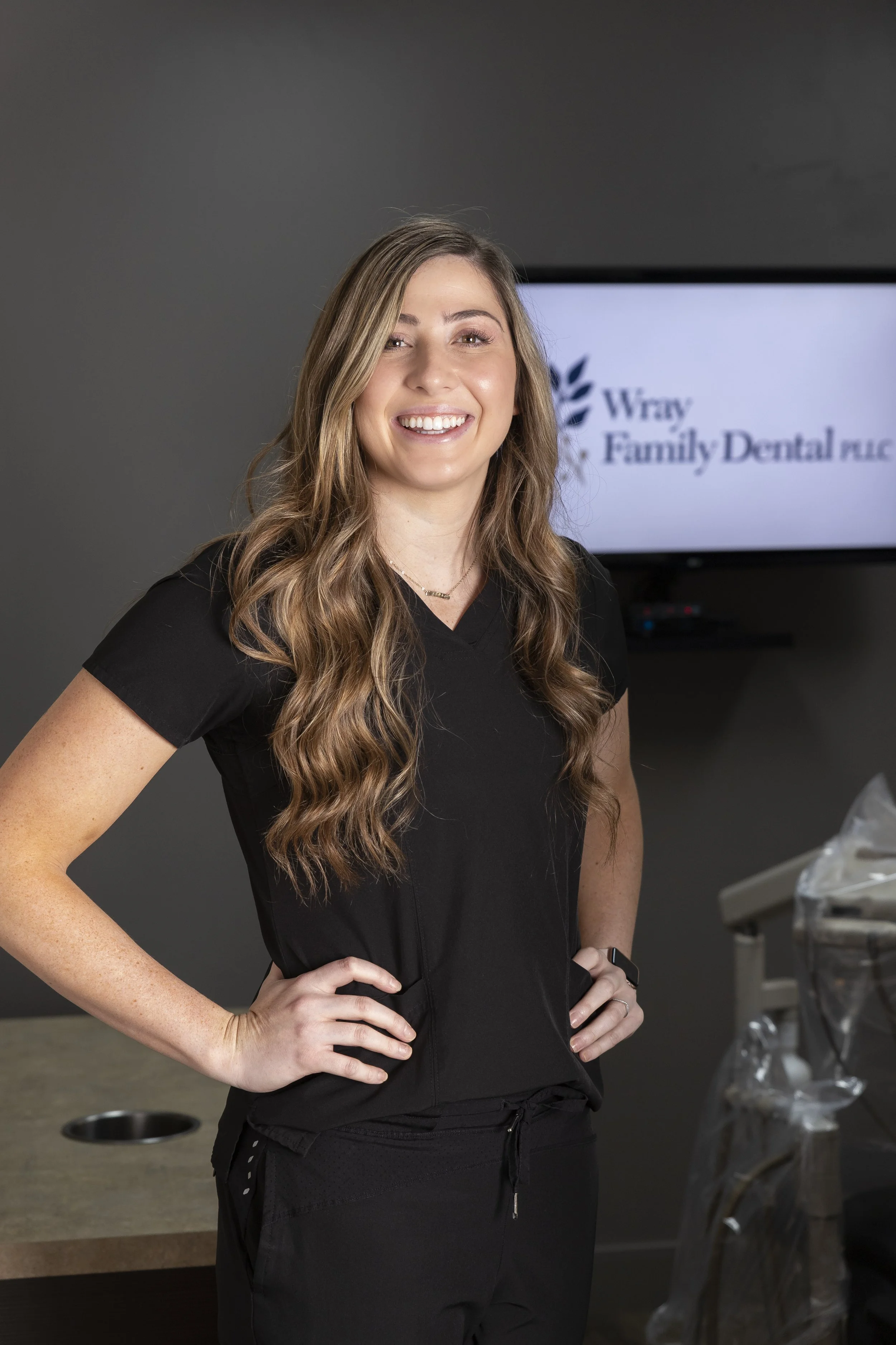 A smiling woman with long brown wavy hair wearing black scrubs stands in a dental office,