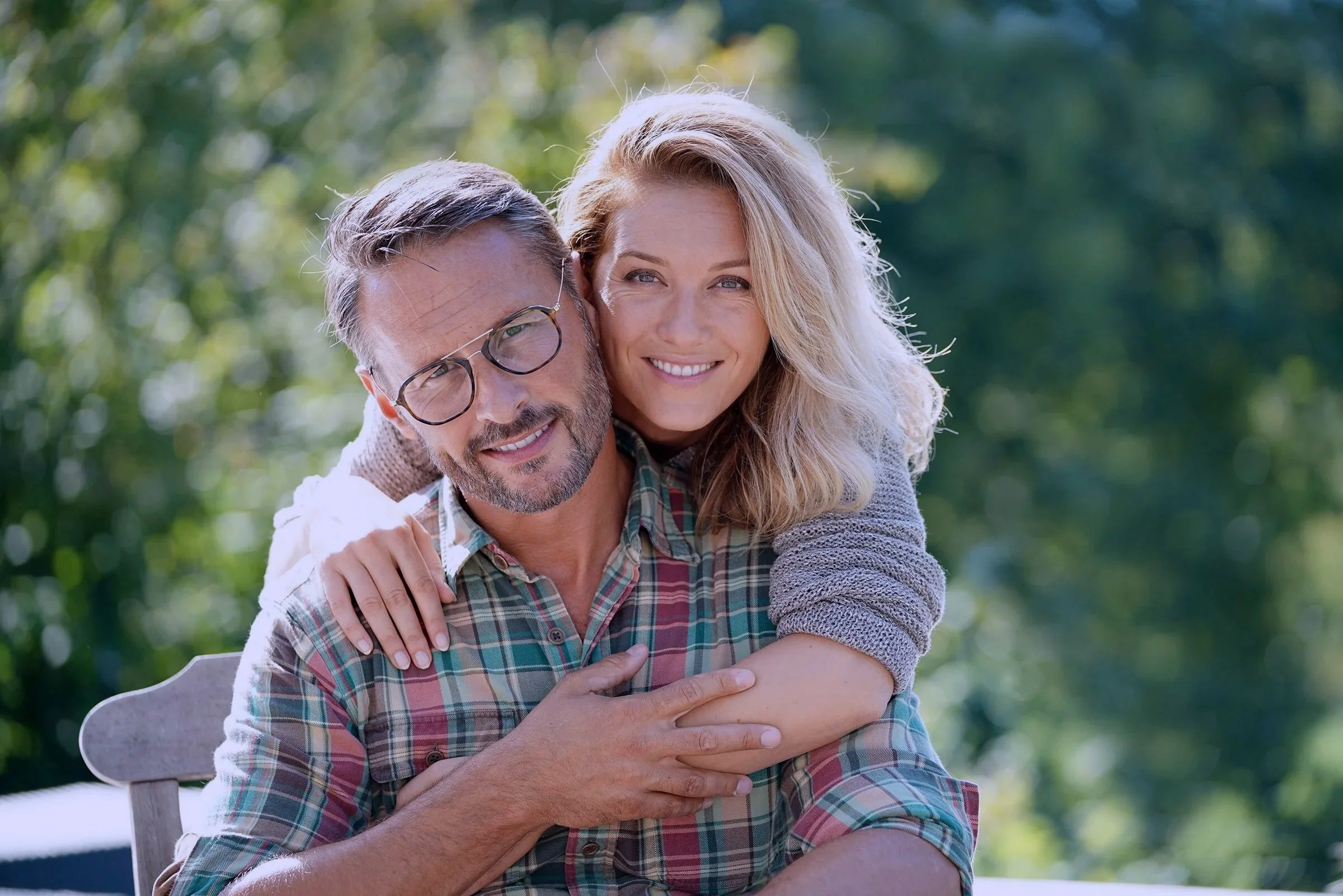 A man with glasses and a woman with blonde hair smiling outdoors, with green trees in the background.