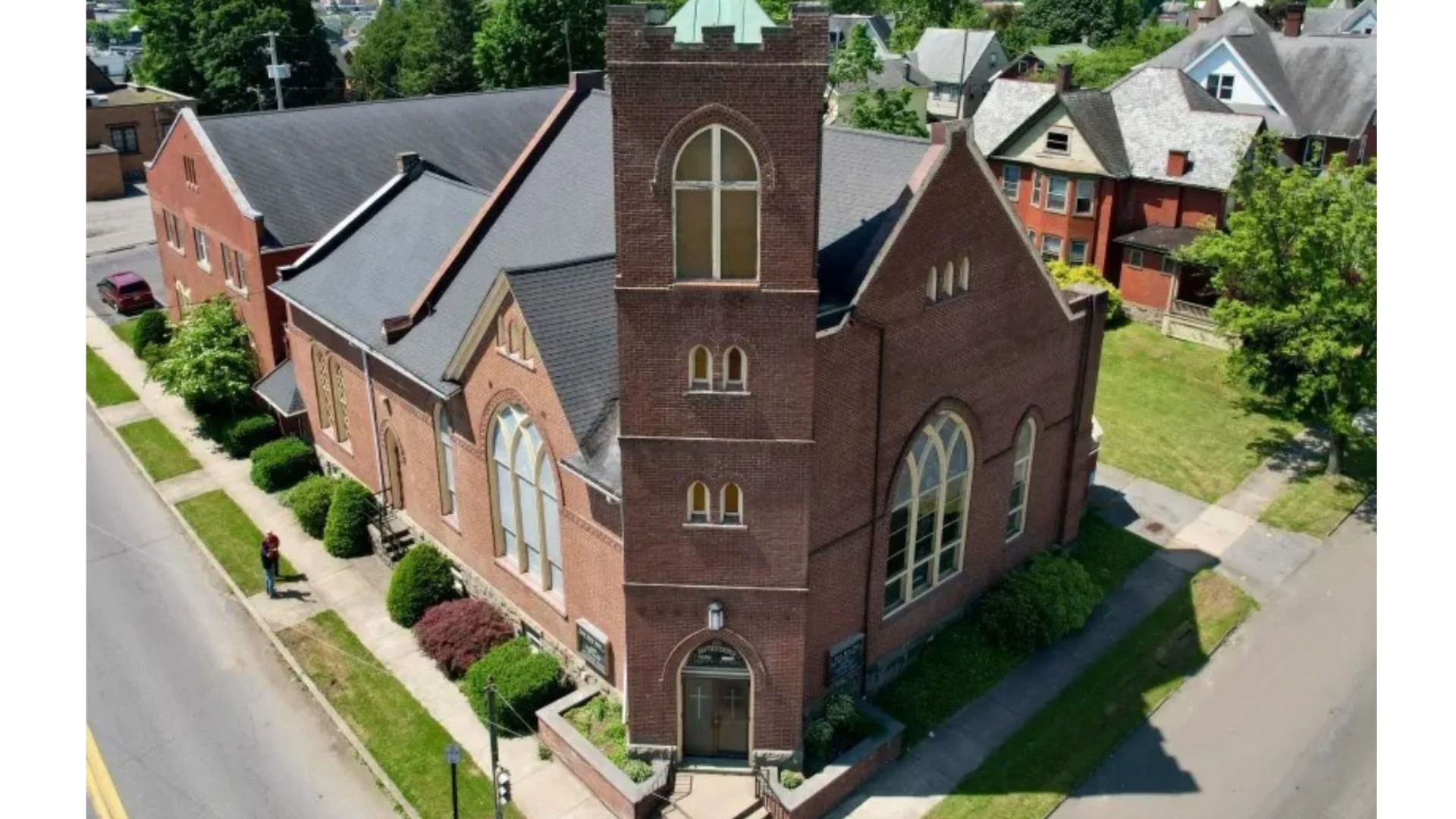 Aerial view of a red brick church with tall pointed windows, a steeple with arched windows, and surrounding residential houses, trees, and sidewalks.