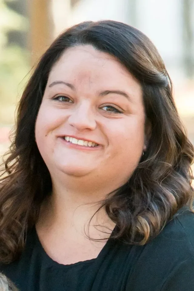 A woman smiling outdoors with brown, wavy hair, wearing a black top.