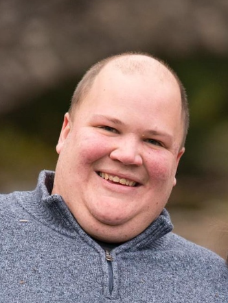 A smiling man outdoors with a blurred natural background.