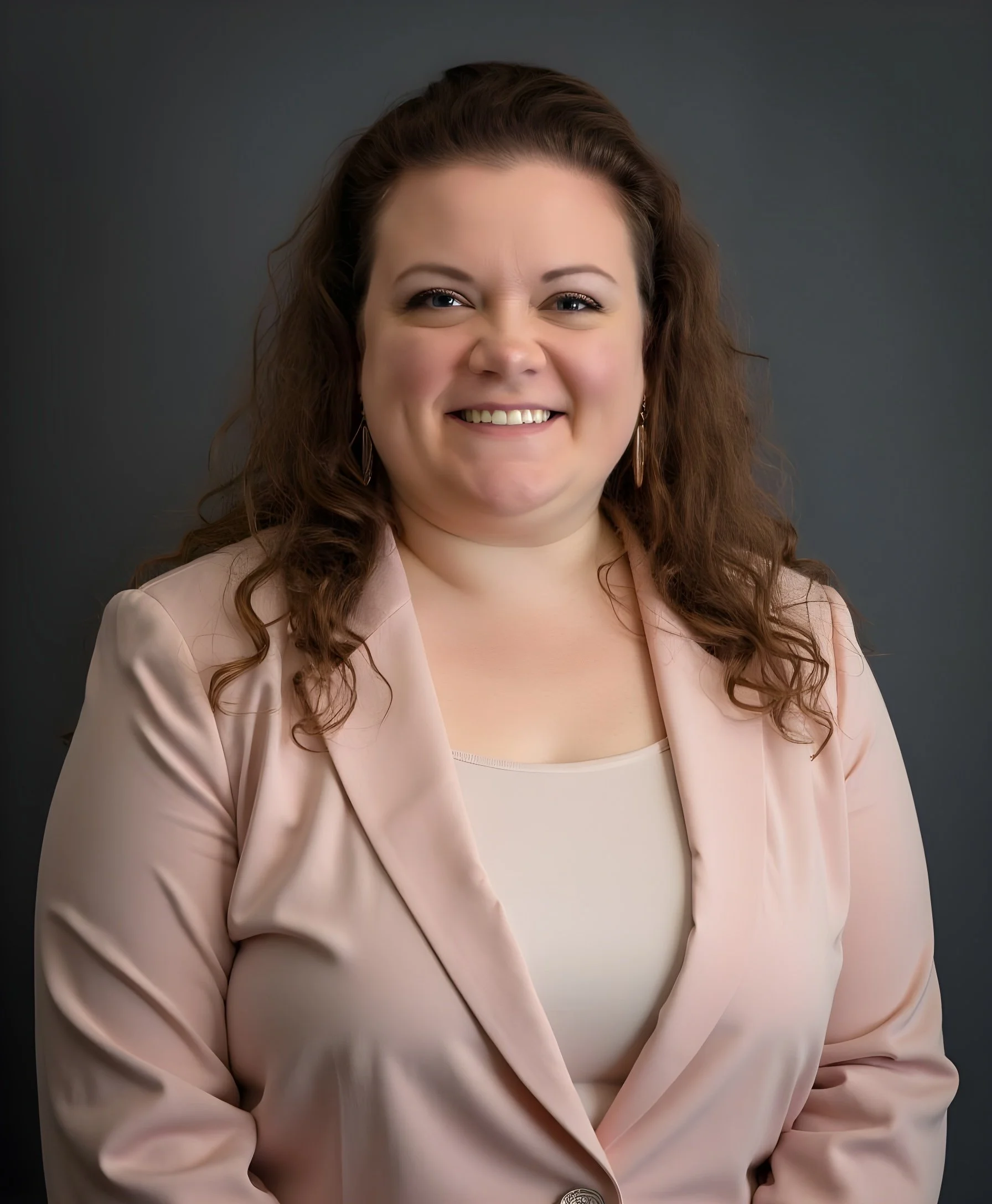 Professional woman with brown, curly hair smiling, wearing a pale pink blazer and a beige top, against a dark gray background.