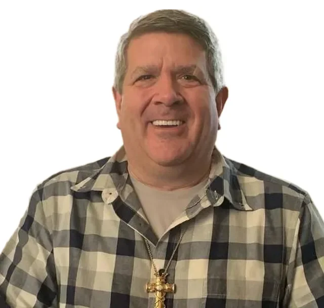 A middle-aged man with gray hair smiling, wearing a plaid shirt and a cross necklace, against a plain white background.