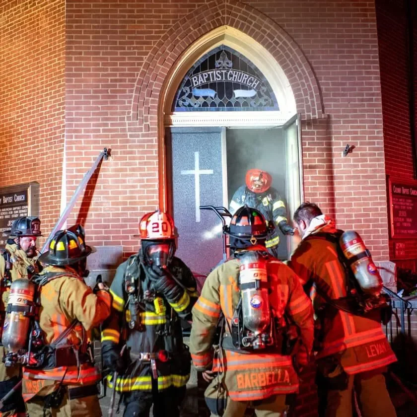 Firefighters in turnout gear and helmets carrying hoses and equipment outside a church with a brick facade and a stained glass window that says 'Baptist Church' and a cross on the door, responding to a fire emergency.