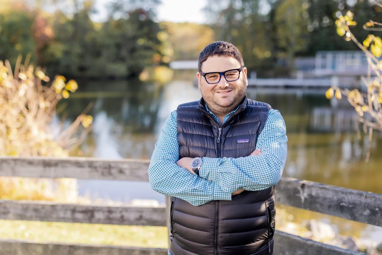 Gilad, with glasses and a short beard standing outdoors by a wooden railing, crossing his arms, wearing a blue checkered shirt and a black puffer vest, with a river and trees in the background during daytime.