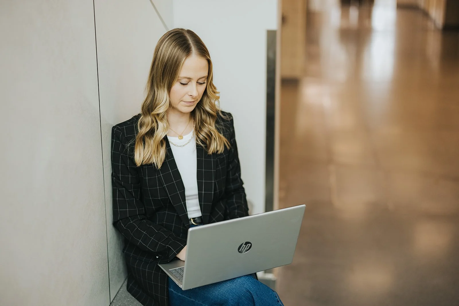 A young woman with wavy blonde hair sitting on the floor, working on a silver HP laptop, dressed in a black checkered blazer and blue jeans, in a modern indoor setting.