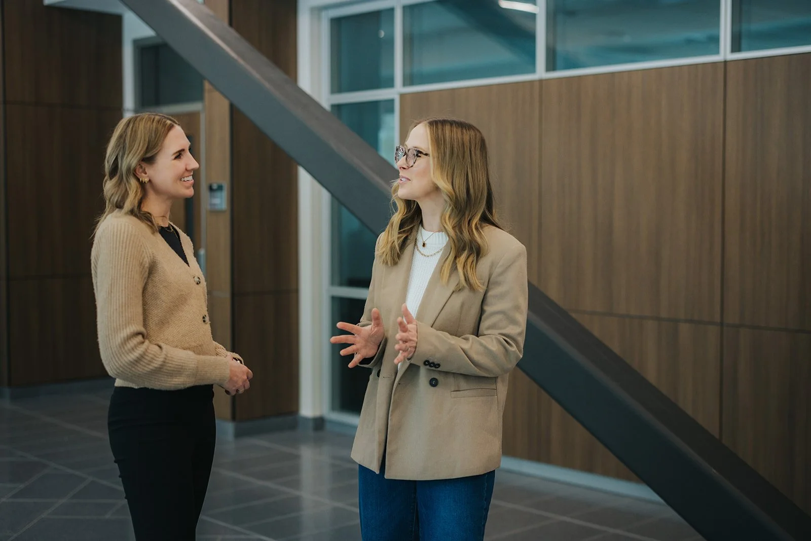 Two women having a conversation in an indoor office setting, with one wearing a beige blazer and the other wearing a tan cardigan.