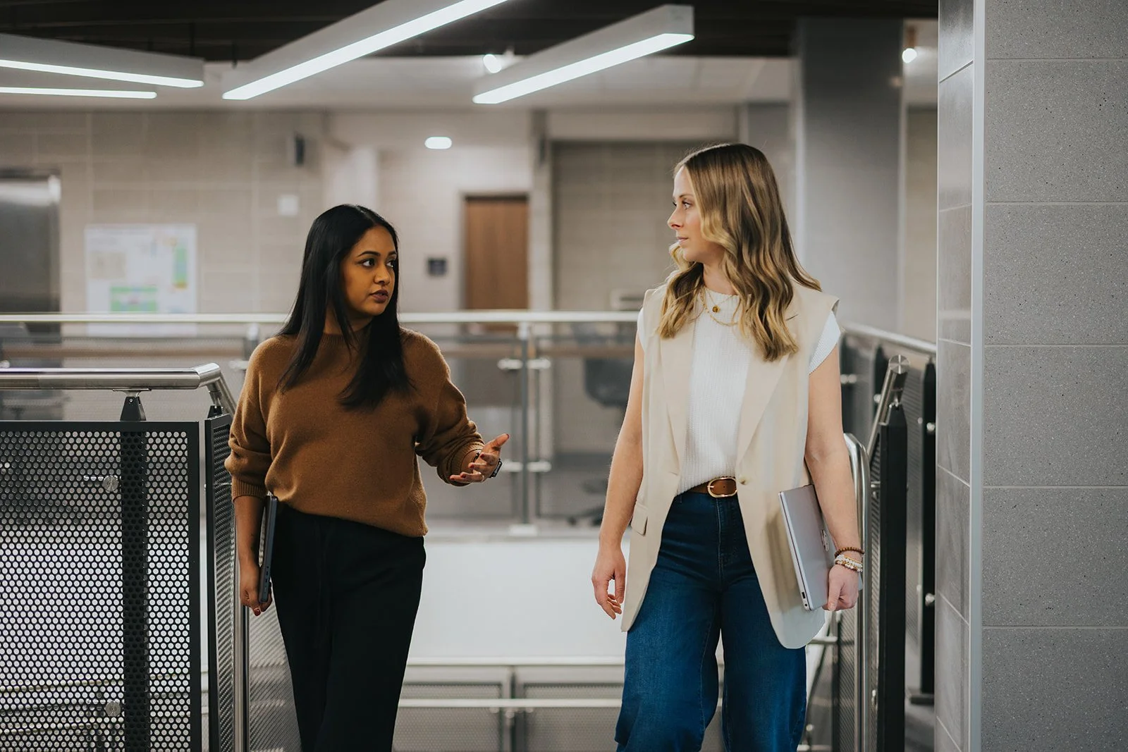 Two women are having a conversation in what appears to be a modern office building or public space. One woman has dark hair and is wearing a brown sweater and black pants, while the other woman has blonde hair and is dressed in a white top and a beige blazer with blue jeans, holding a laptop.