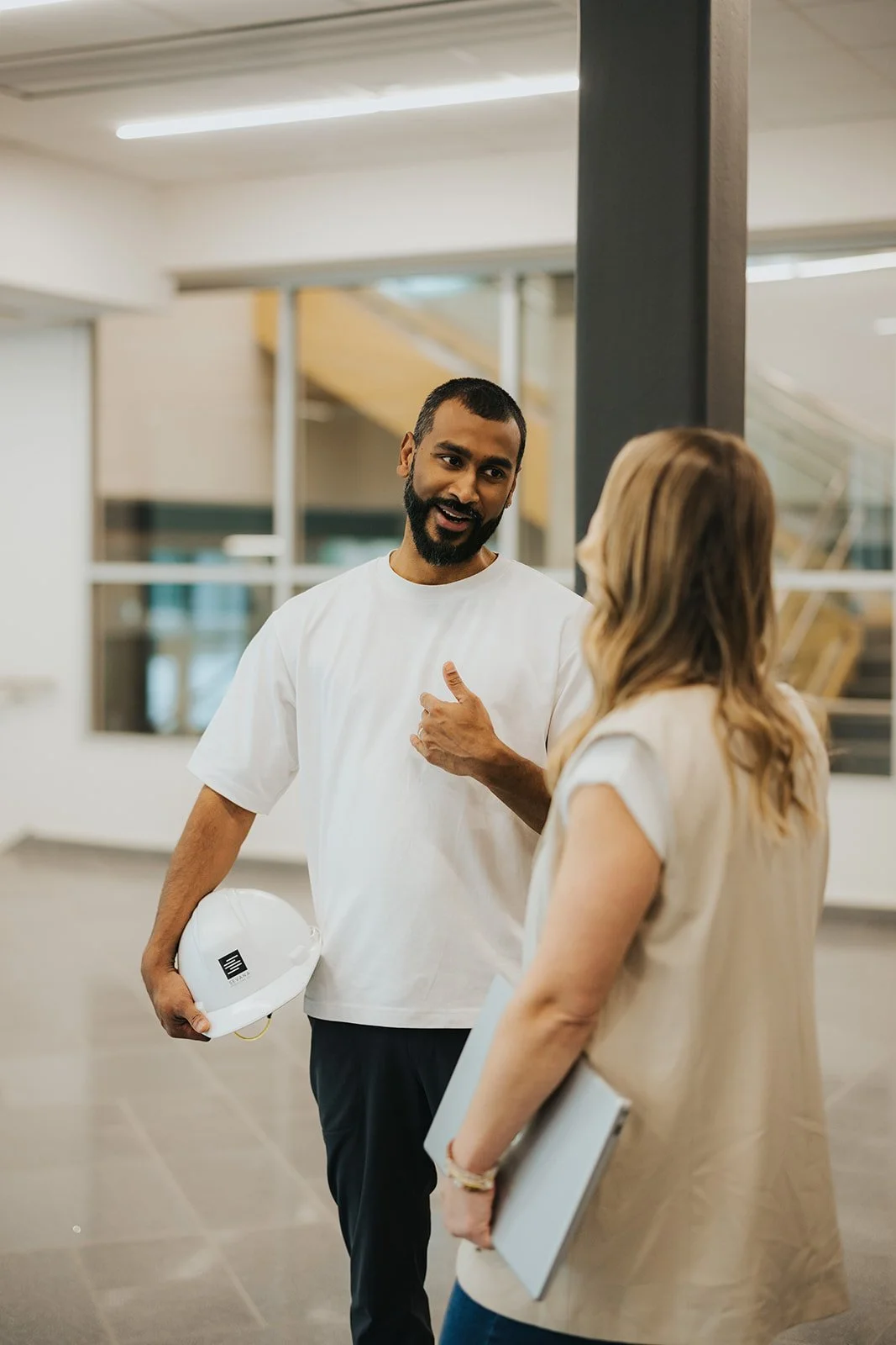 Two people having a conversation in a modern office building. The man, with a beard and wearing a white t-shirt, holds a white safety helmet. The woman, with blonde hair, wears a beige vest and holds a tablet.