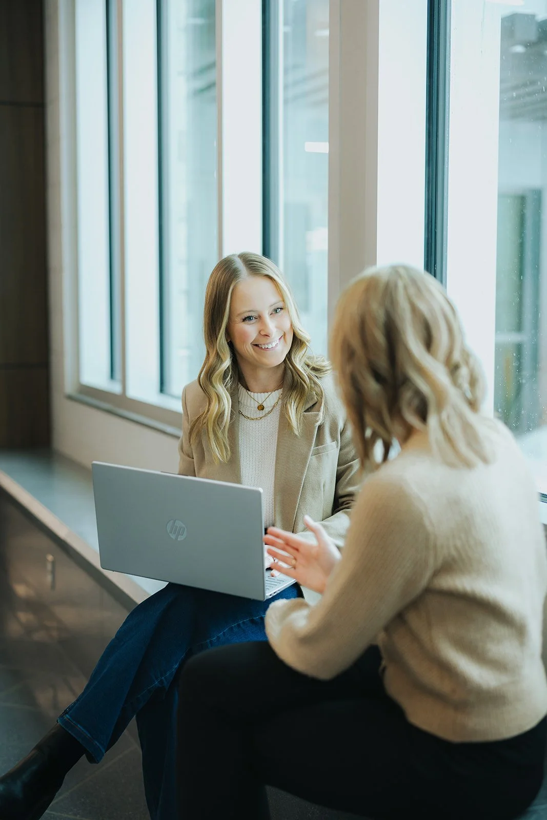 Two women sitting by a large window, engaged in conversation with one woman smiling while holding a laptop on her lap.