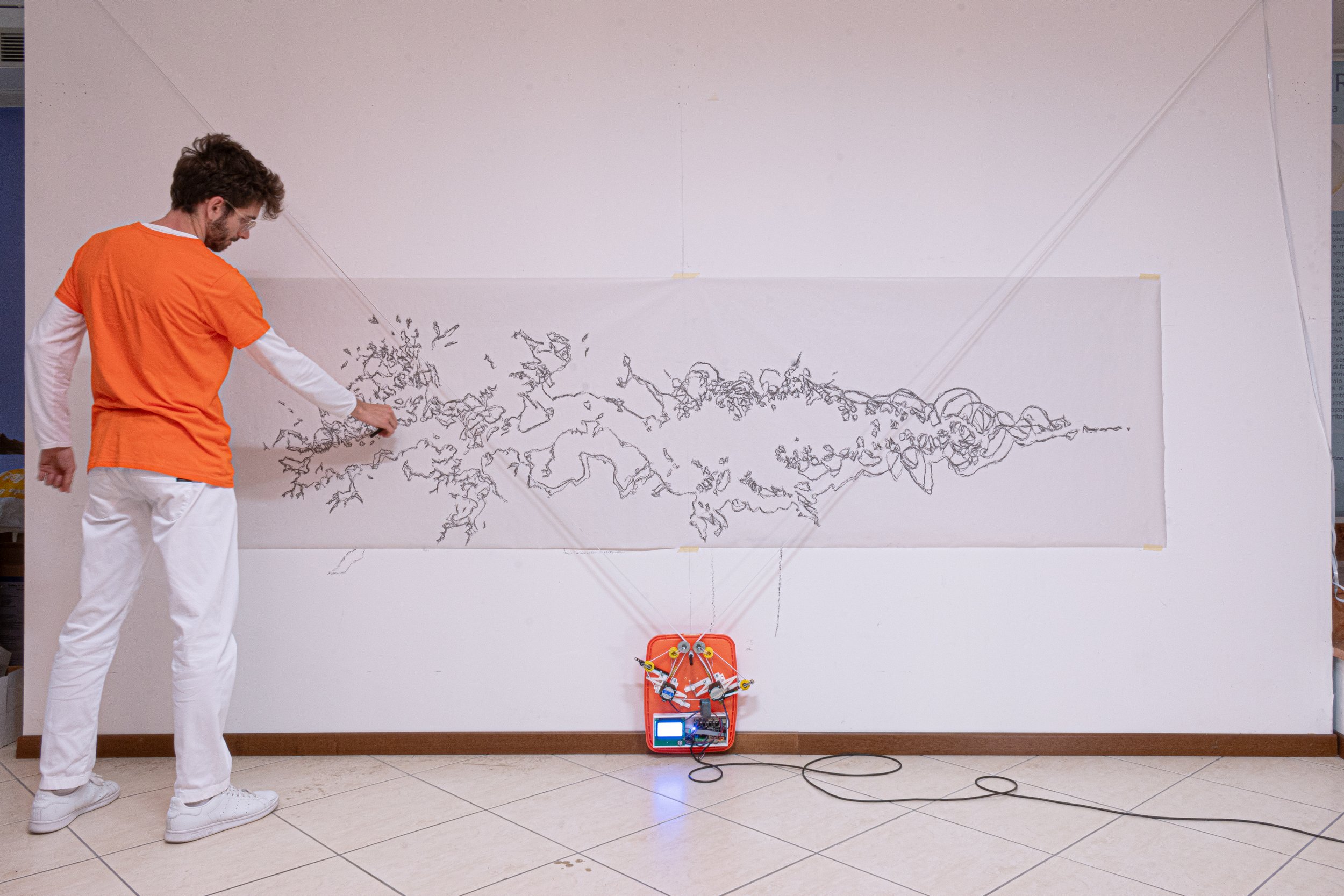 Un uomo con maglietta arancione e pantaloni bianchi sta dipingendo a mano libera una linea irregolare su un grande pannello bianco, collegato a un dispositivo elettronico di colore arancione sul pavimento.