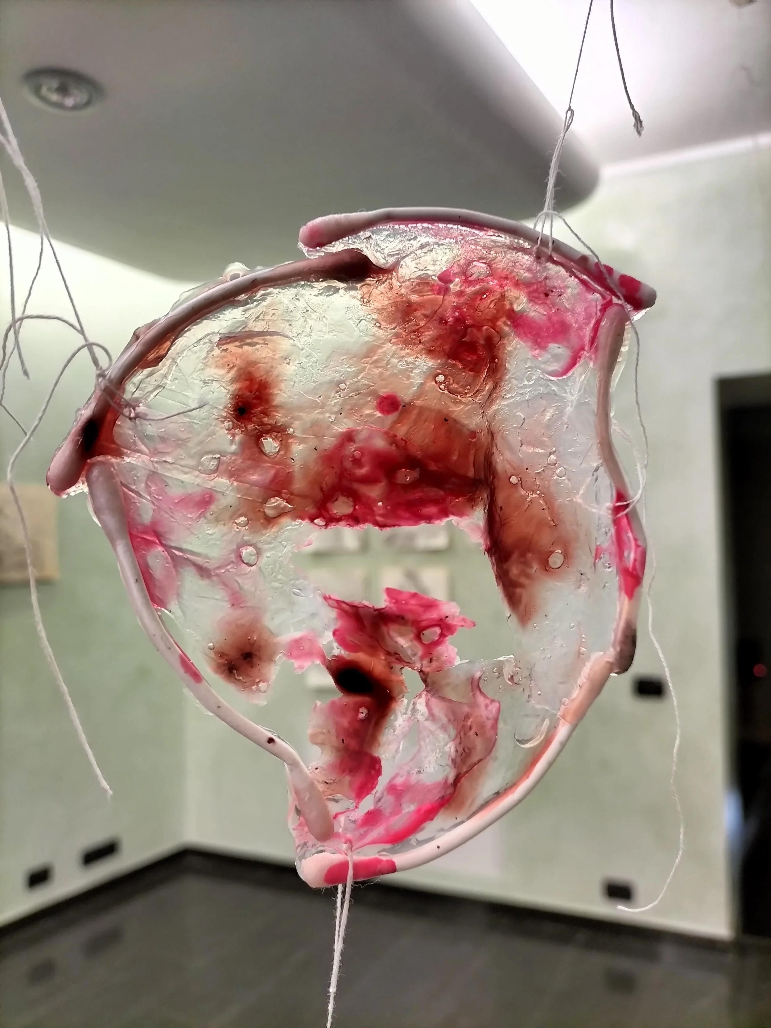 Un'opera d'arte astratta composta da vetro trasparente con macchie e strisce di colore rosso, rosa e marrone, sospesa con fili sottili in un ambiente interno.