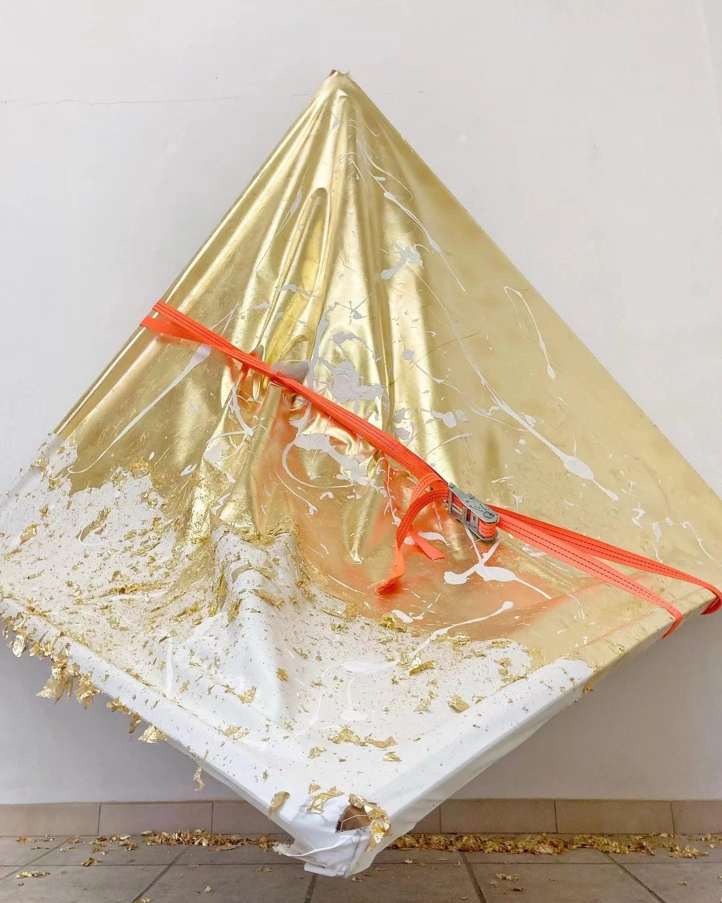 Installazione artistica con una struttura a forma di piramide avvolta in materiale dorato e bianco, legata con una cinghia arancione.