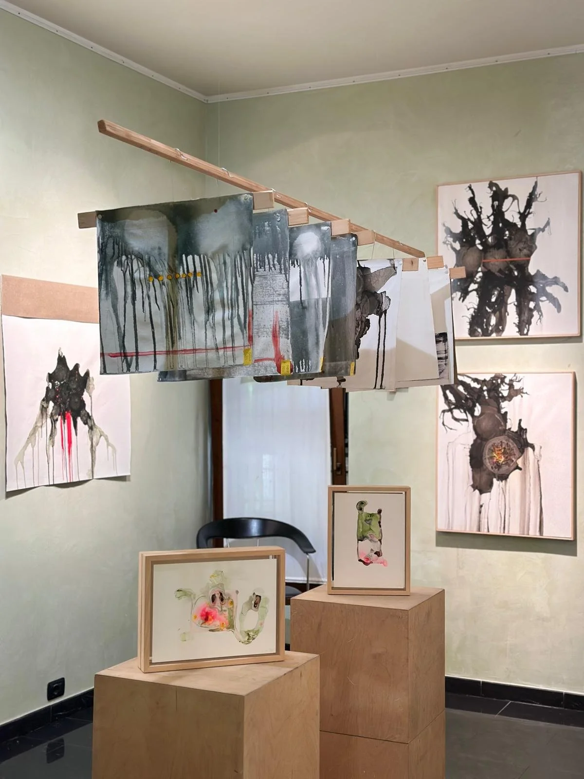 Opere d'arte astratte e dipinti in una mostra o galleria, con dipinti di colori e forme astratte appesi alle pareti e alcune opere più piccole in cornice posizionate su un piedistallo di legno.