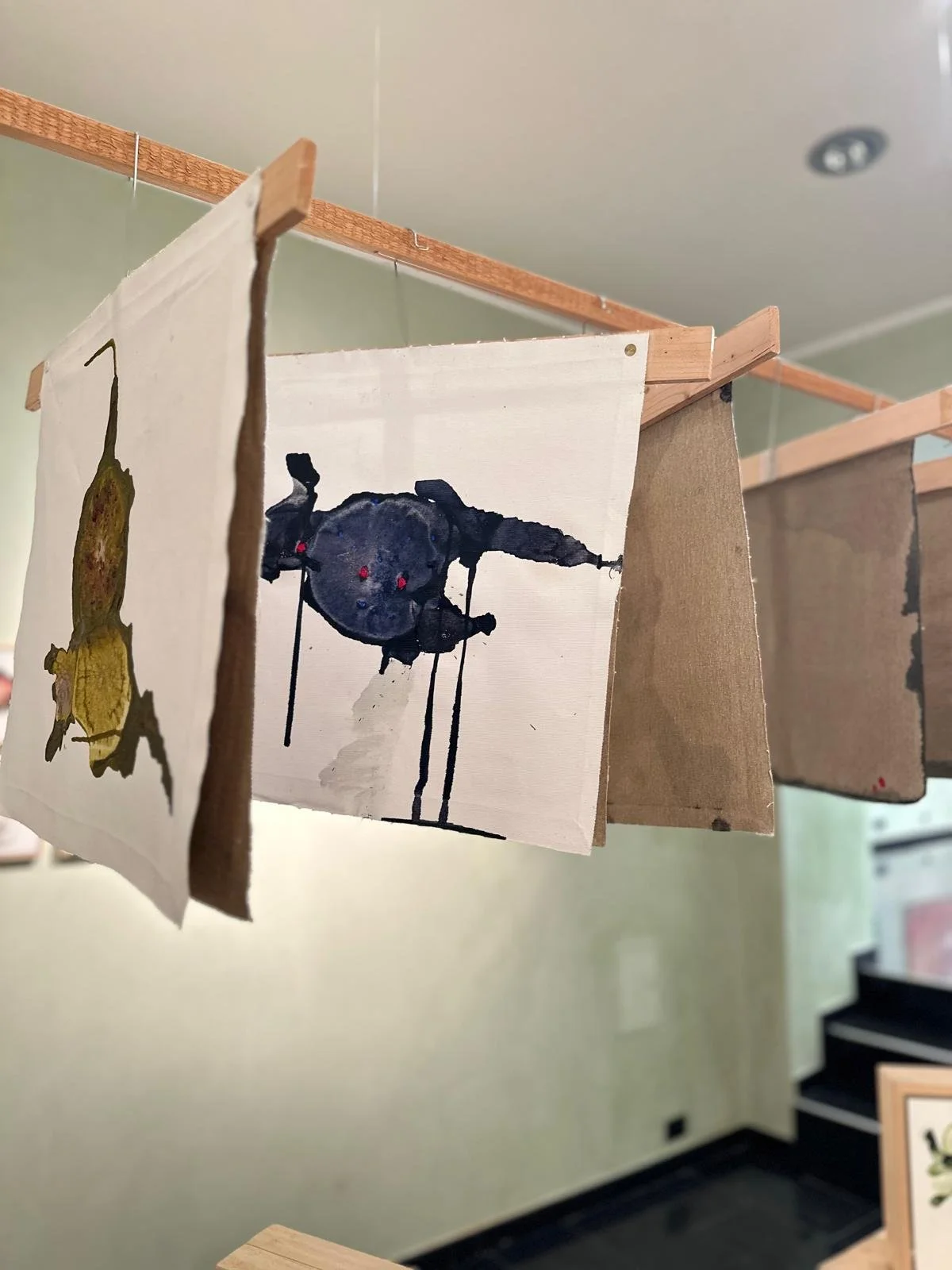 Opere d'arte su tessuto appese su fili da un'asta di legno, con pareti verdi sullo sfondo.