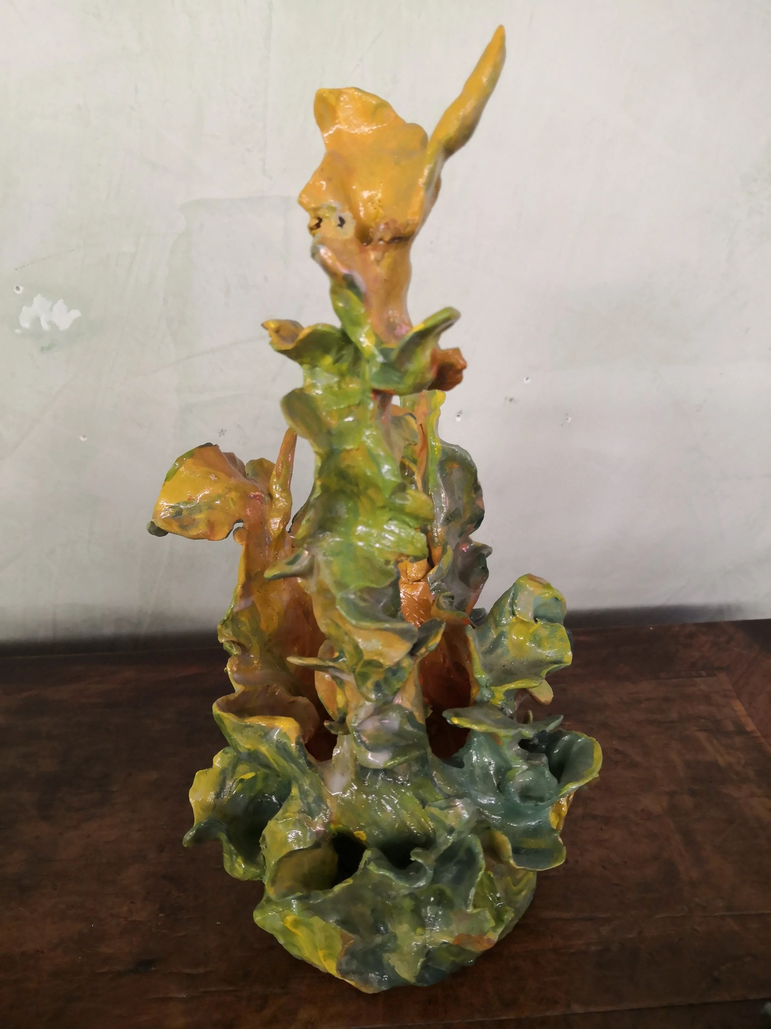 Statua in ceramica con forma di foglie e rami, di colore verde, marrone e giallo, posizionata su un tavolo di legno.