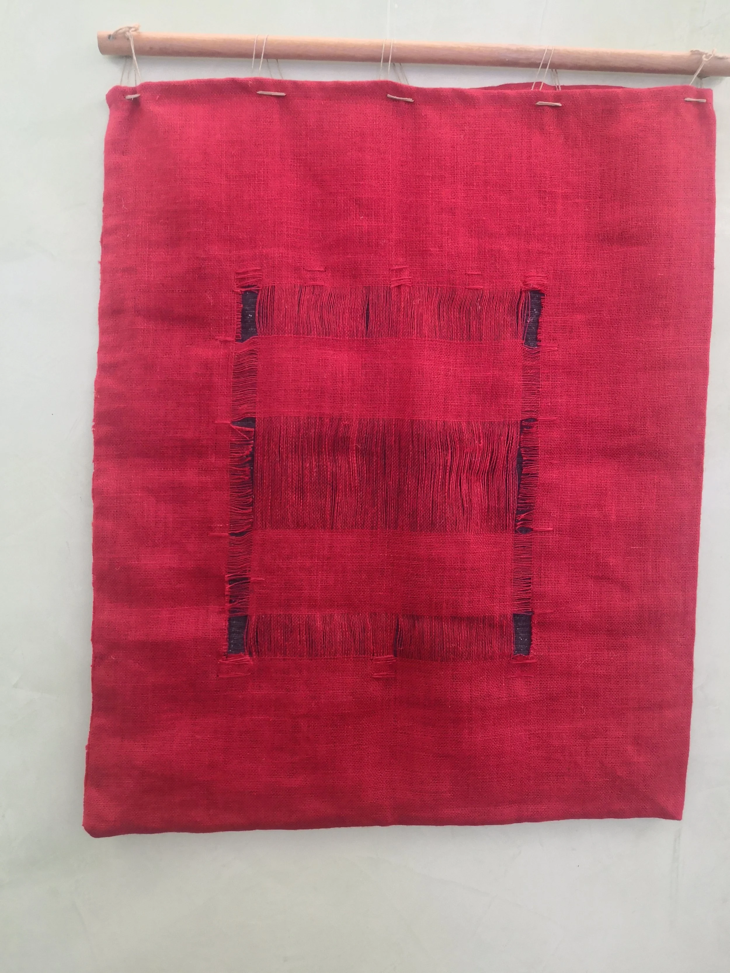 Tessuto di colore rosso appeso a un'asta di legno. Il tessuto presenta una nervatura centrale e un motivo geometrico elaborato con diverse tonalità di rosso e linee nere.