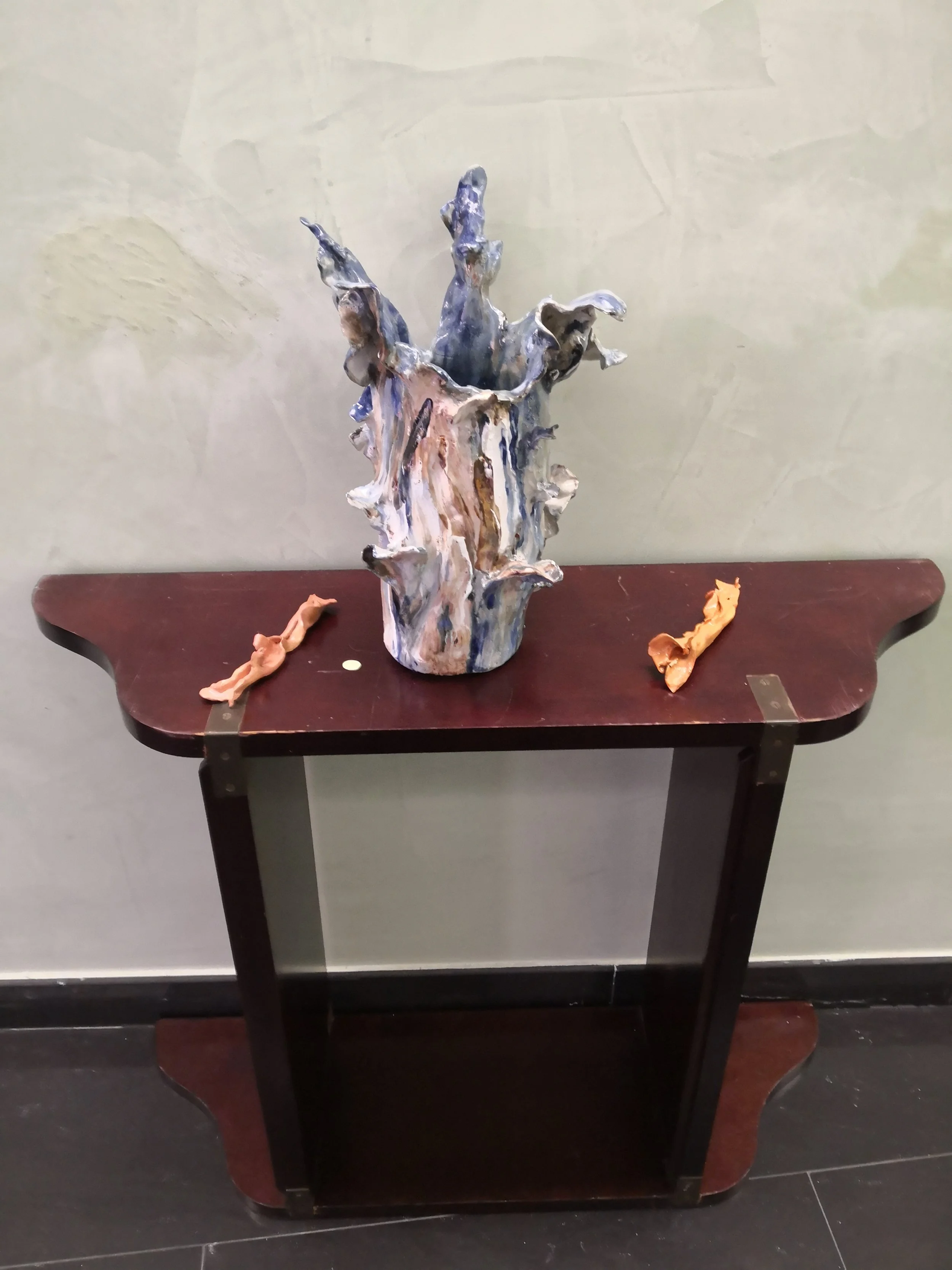 Una consolle di legno scuro con un oggetto decorativo al centro, che sembra un pezzo di arte astratta con colori blu, bianco e marrone, e due pezzi di creta o pasta modellabile ai lati.