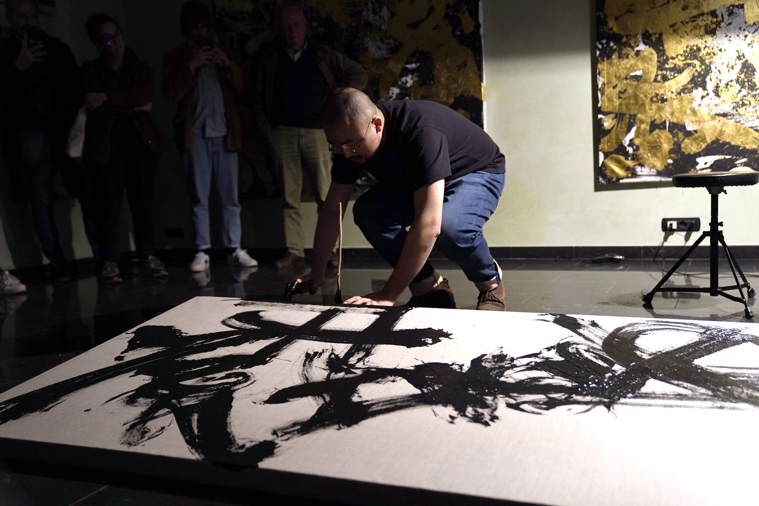 Artista mentre dipinge un quadro grande con pennellate nere su una tela, circondato da un pubblico.