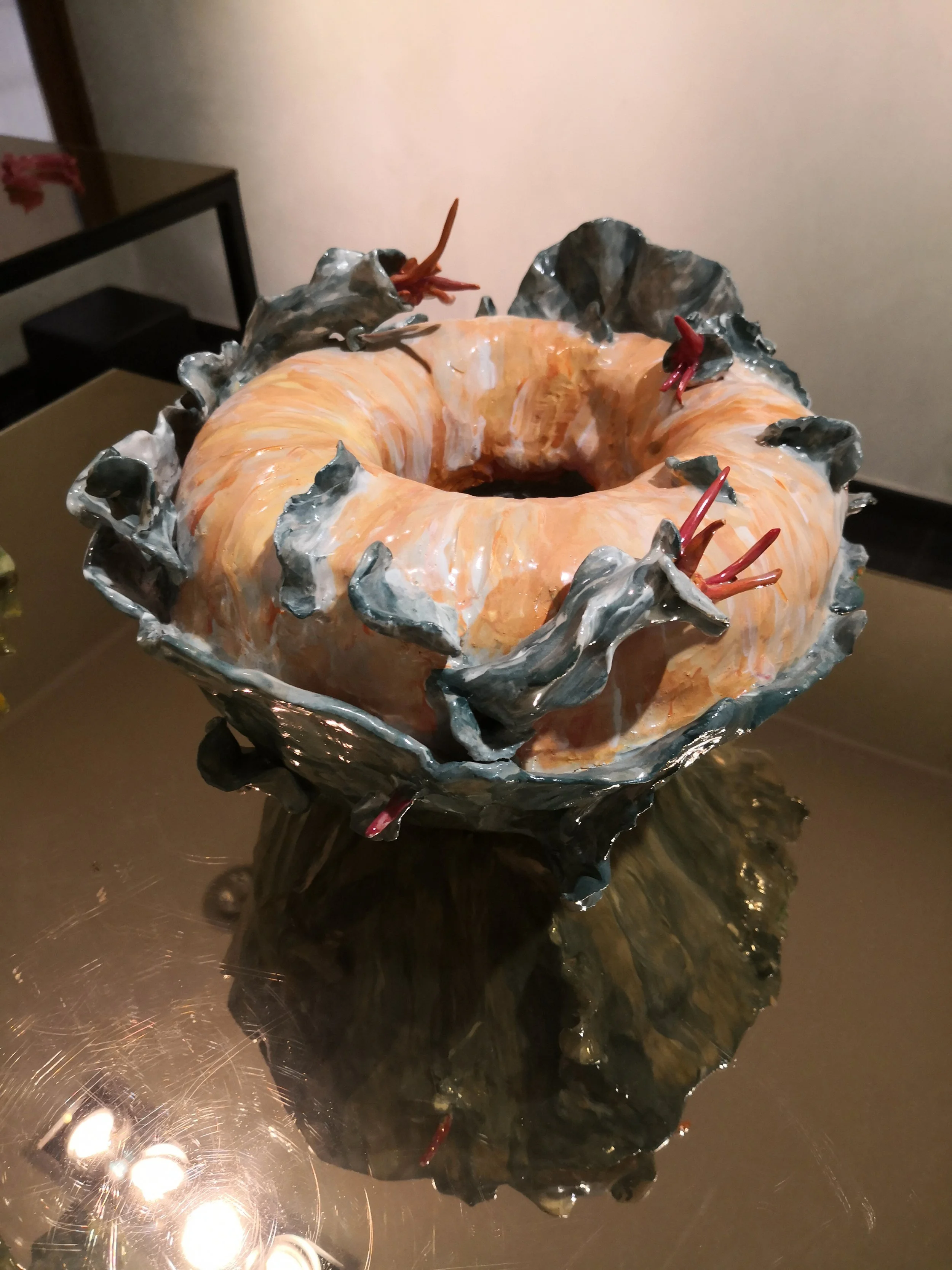 Una scultura artistica che somiglia a una ciambella con elementi colorati e metallici, posizionata su un tavolo riflettente.