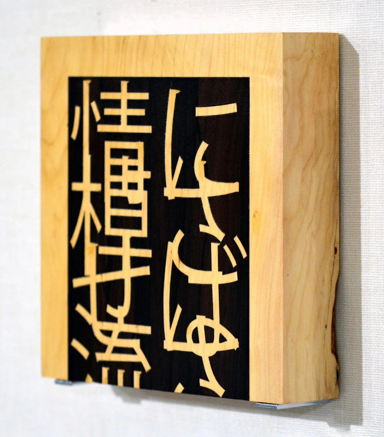 Pannello di legno con scritte giapponesi in rilievo nere su sfondo bianco, con cornice di legno naturale.