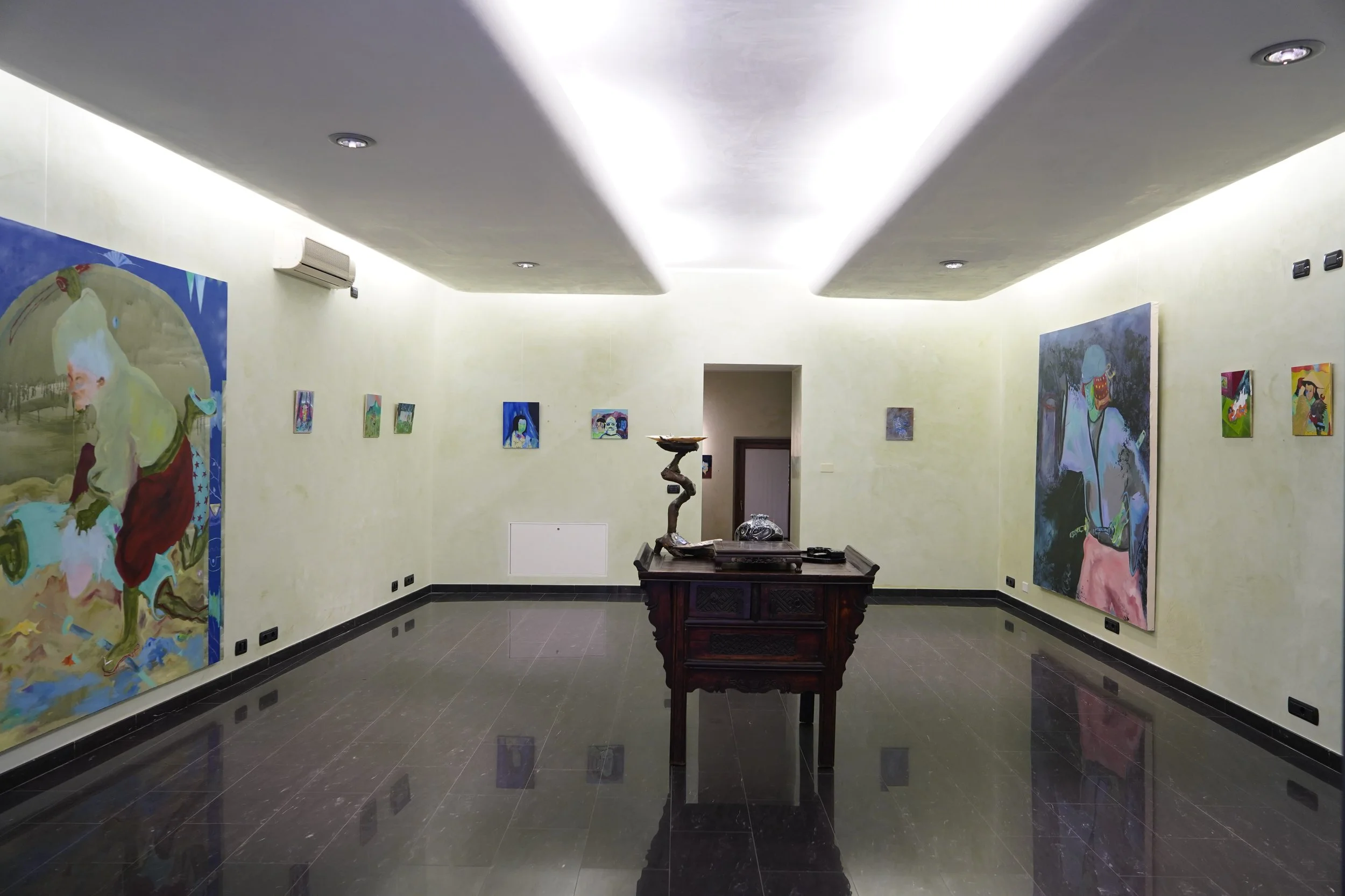 Una sala espositiva con pareti di colore chiaro, quadri di arte moderna appesi e un tavolo centrale con sculture e oggetti artistici.