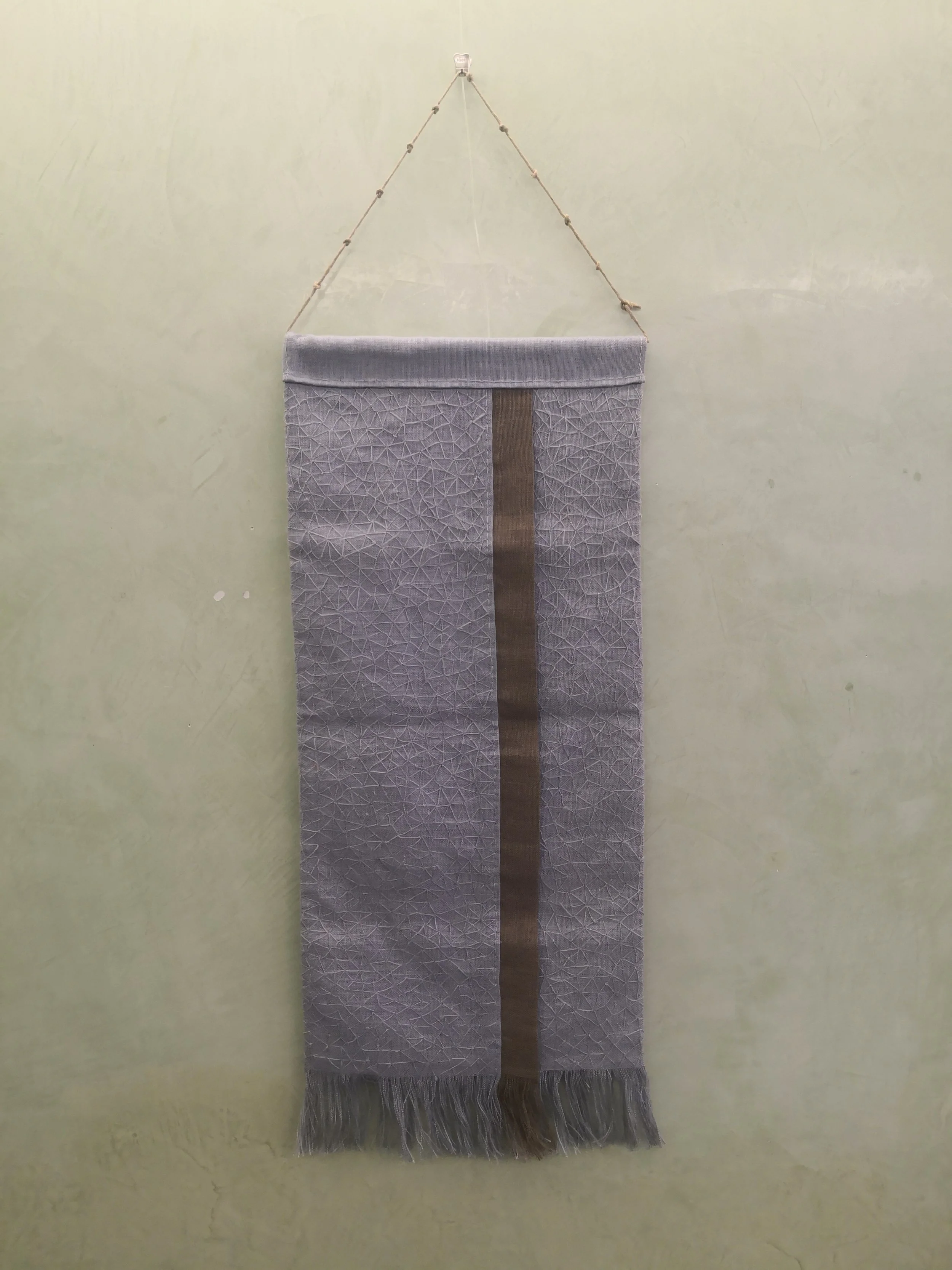 Tessuto decorativo grigio con motivo geometrico e una fascia centrale marrone, appeso a parete con una catena e frange in basso.