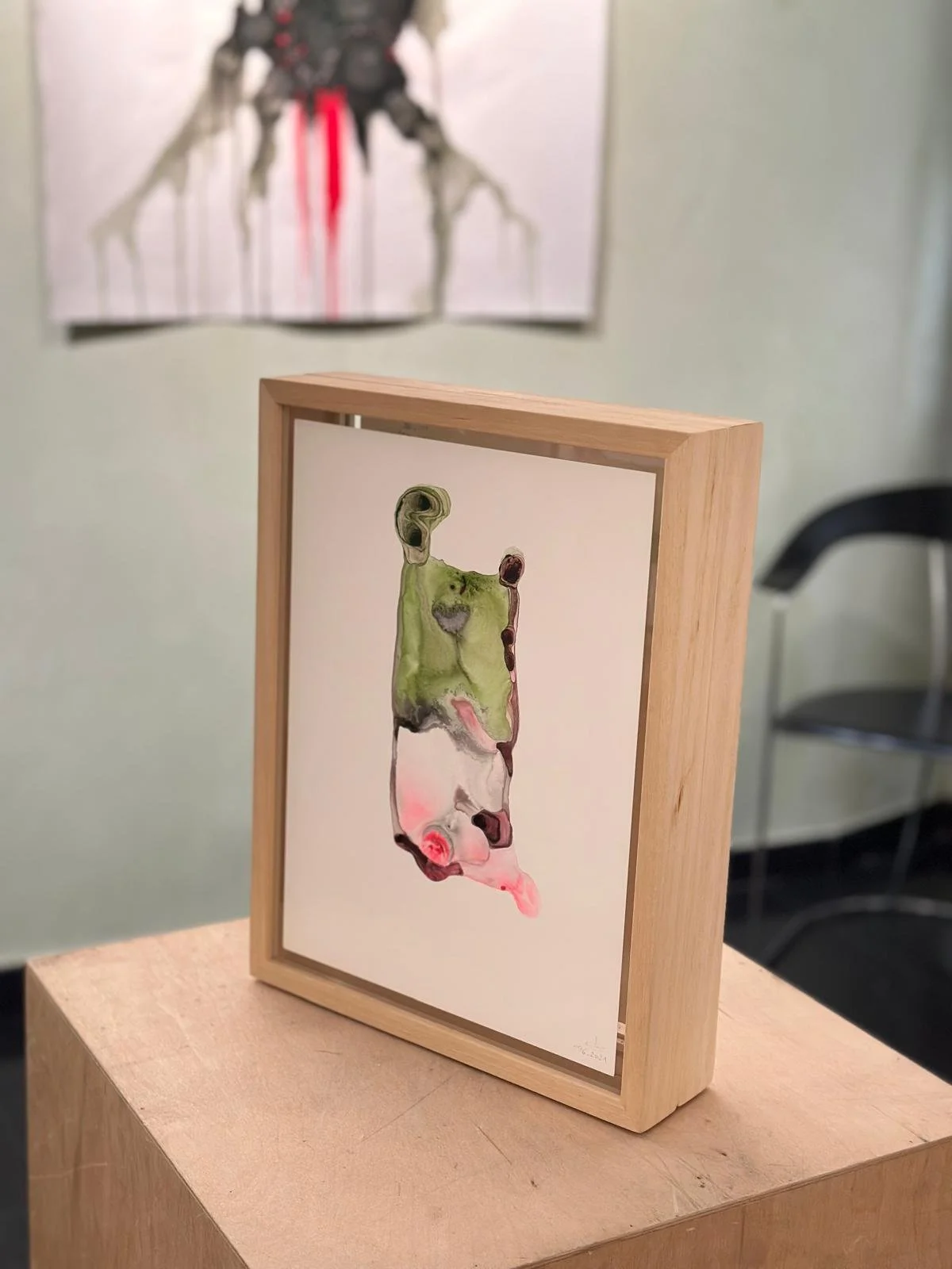 Un quadro astratto con forme fluide in colori verdi, rosa, bianco e nero, incastonato in una cornice di legno, posizionato su un supporto di legno. Sullo sfondo, un'altra opera d'arte è appesa al muro, con colori più neutri e dettagli più distinti.