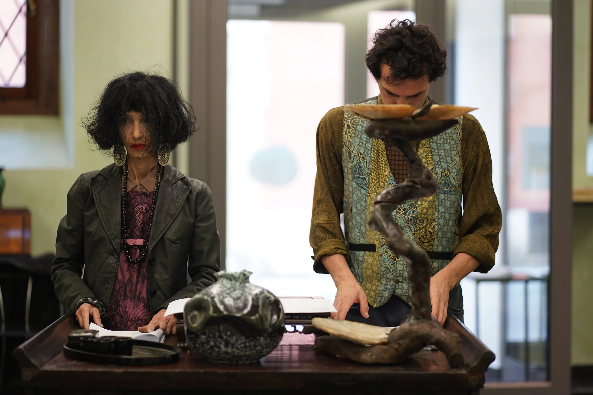 Due persone alla mostra, una con vestiti colorati e una con cappelliere e vestiti scuri, osservano oggetti d'arte e sculture su un tavolo.