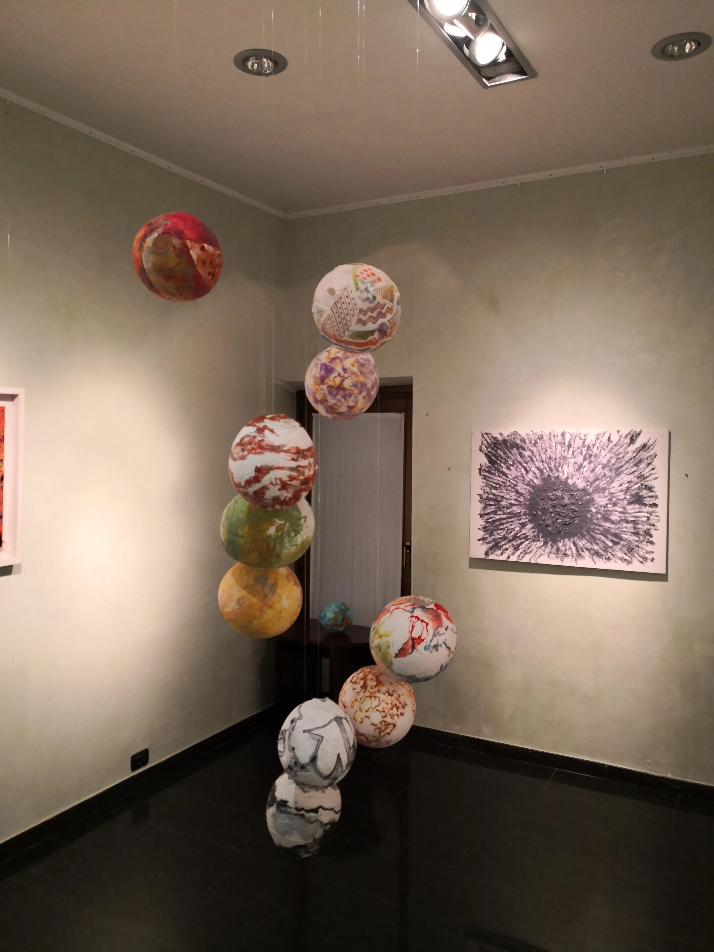 Installazione artistica con dieci sfere sospese, decorate con motivi astratti e colori vivaci, in una stanza con pareti verdi e un quadro astratto appeso.