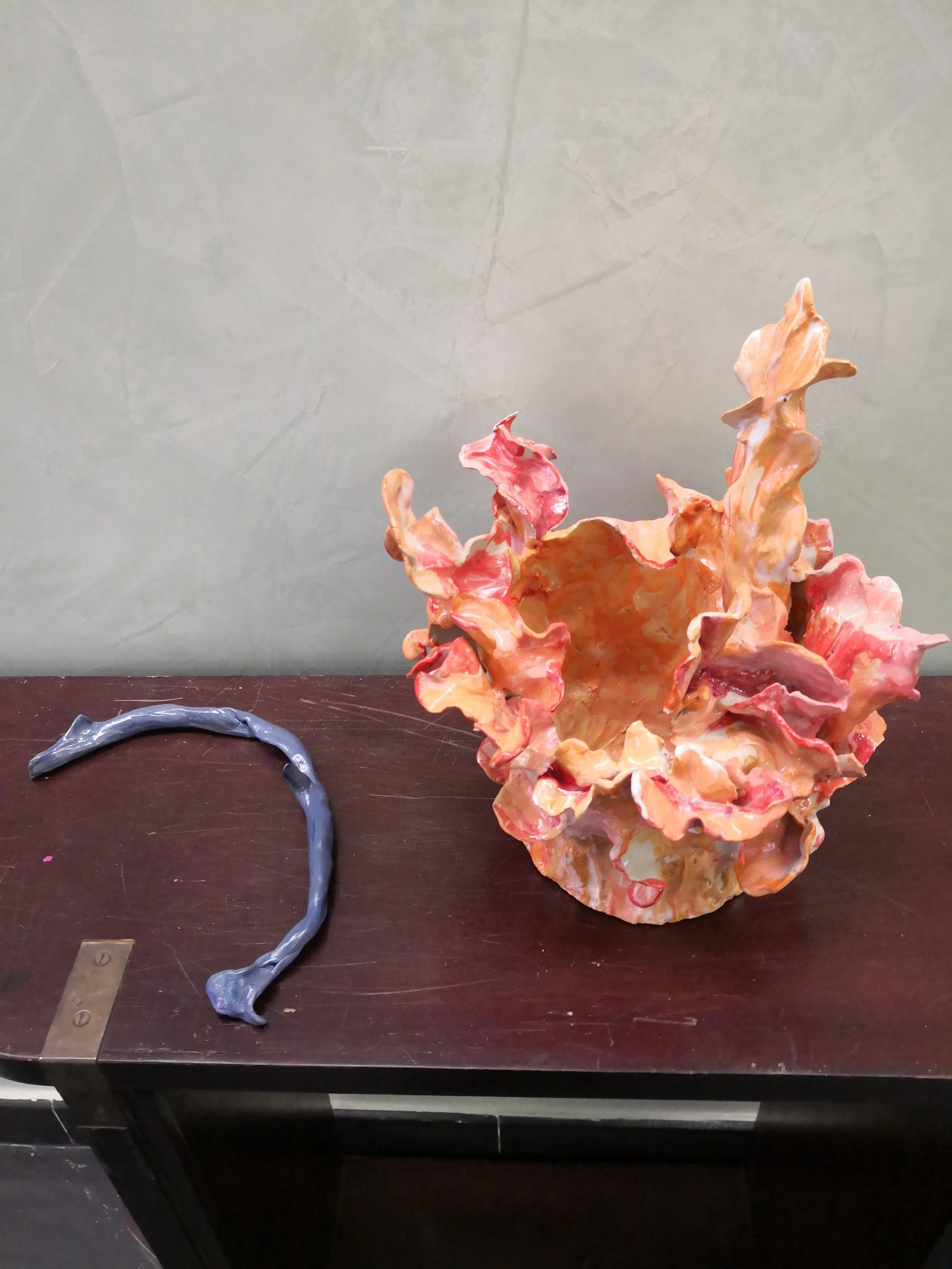 Scultura artistica colorata di forma organica con creste e incavi, collocata su un tavolo di legno scuro con parete grigia sullo sfondo. Accanto, un oggetto blu a forma di fune o cavo.