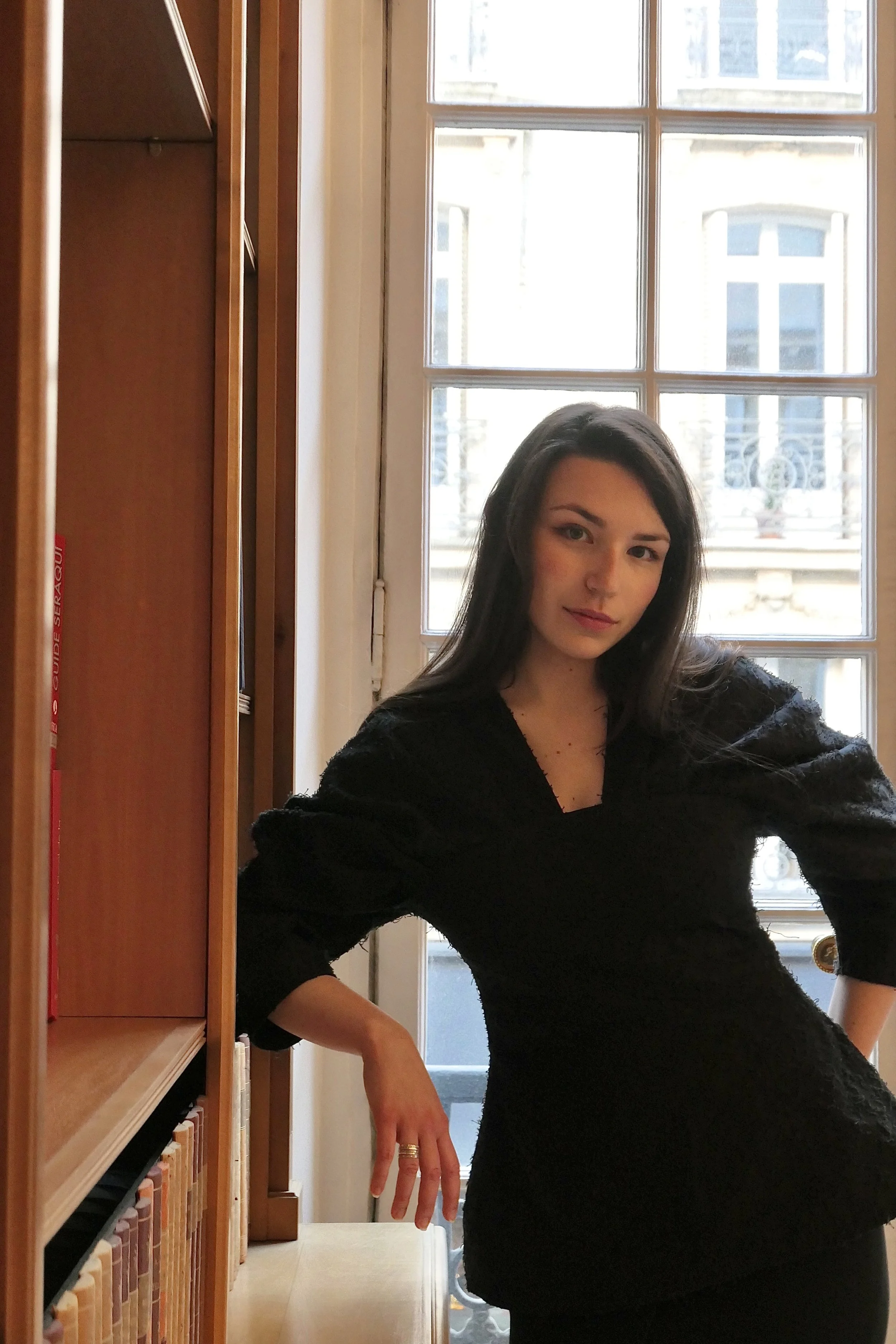 Une jeune femme pose devant une grande fenêtre avec des livres sur une étagère à sa gauche, à l'intérieur d'une pièce.