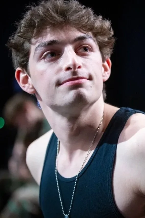 Close-up of a young man with wavy brown hair, wearing a dark tank top and a silver chain necklace, looking slightly up and to the right in a dimly lit background.