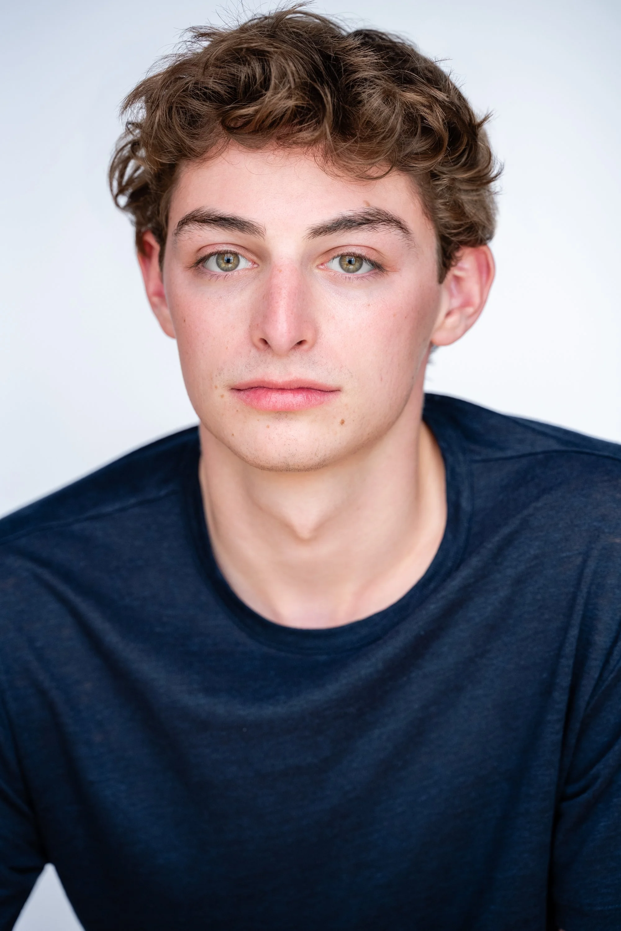 A young man with short, curly brown hair, light skin, and green eyes, wearing a dark blue shirt, looking directly at the camera against a light background.