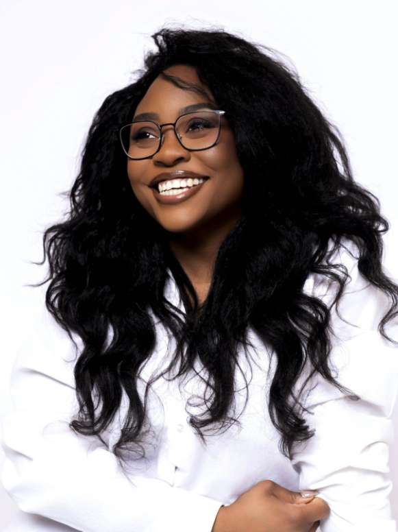 A woman with long, curly black hair and glasses smiling, wearing a white shirt, against a plain white background.