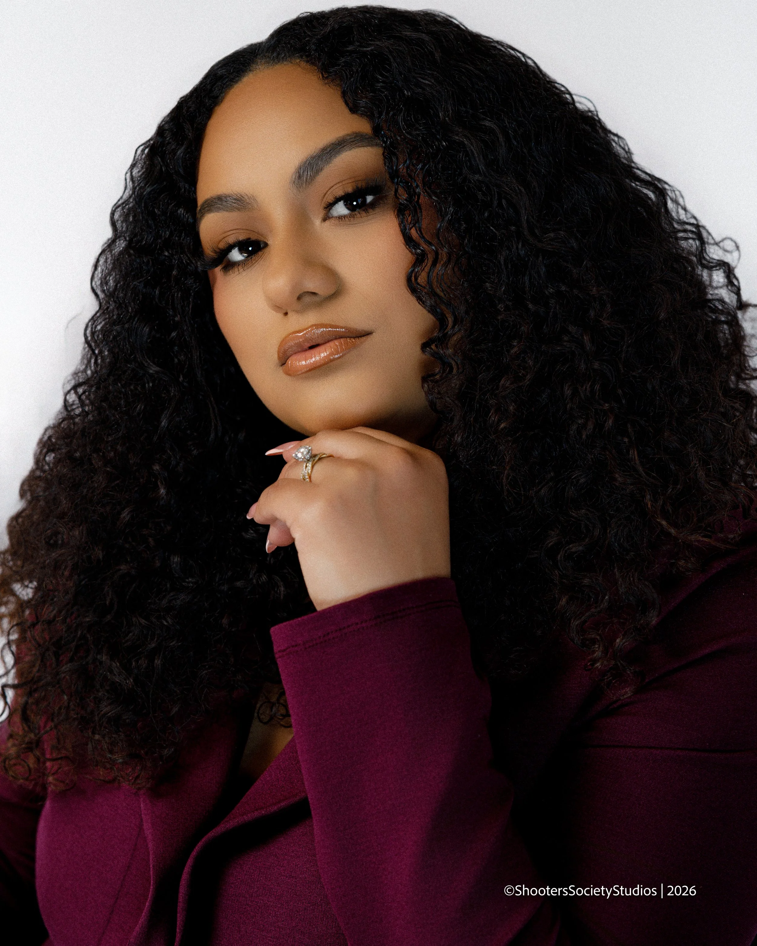 A woman with curly black hair, wearing a maroon top, resting her chin on her hand with a ring visible, posing against a plain white background.