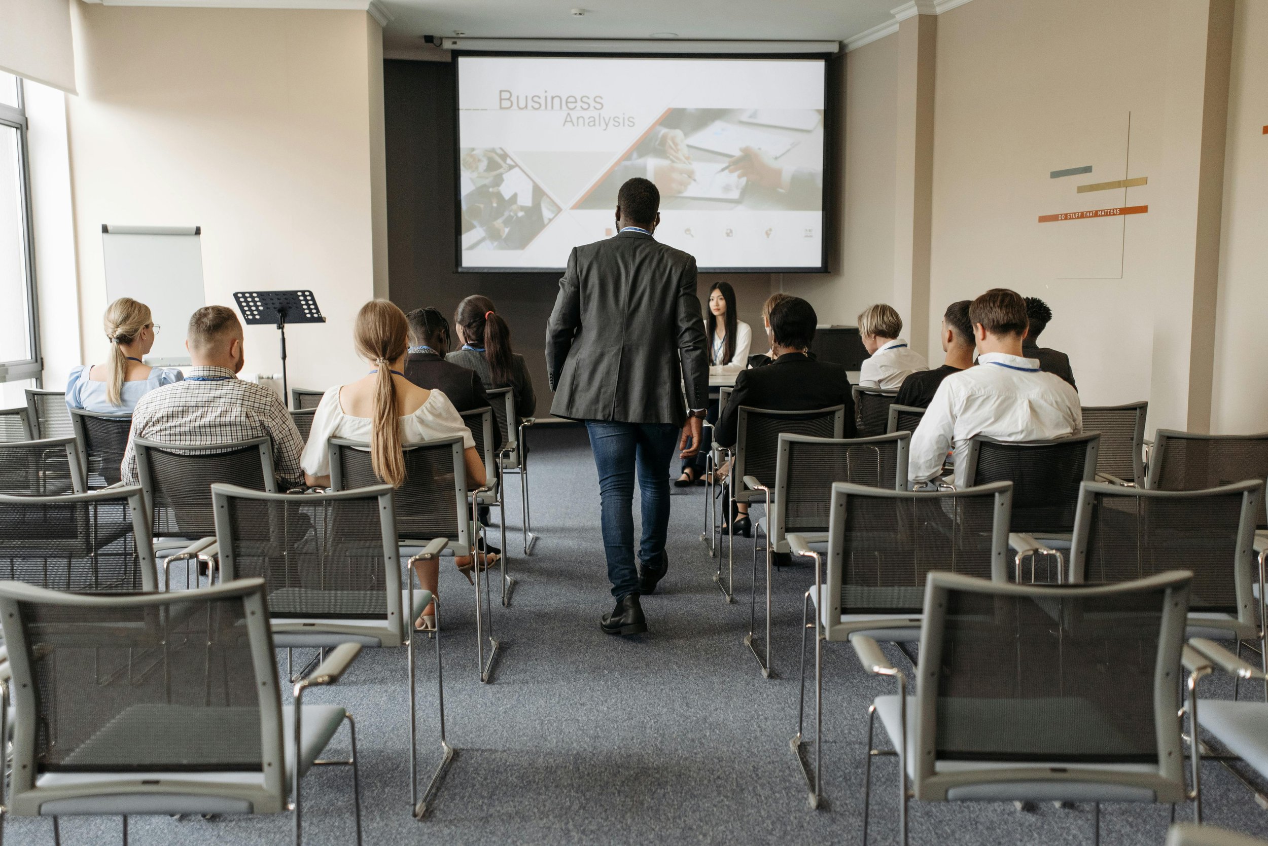 A business meeting classroom with attendees seated facing a presentation screen displaying 'Business Analysis' and a woman walking down the aisle.